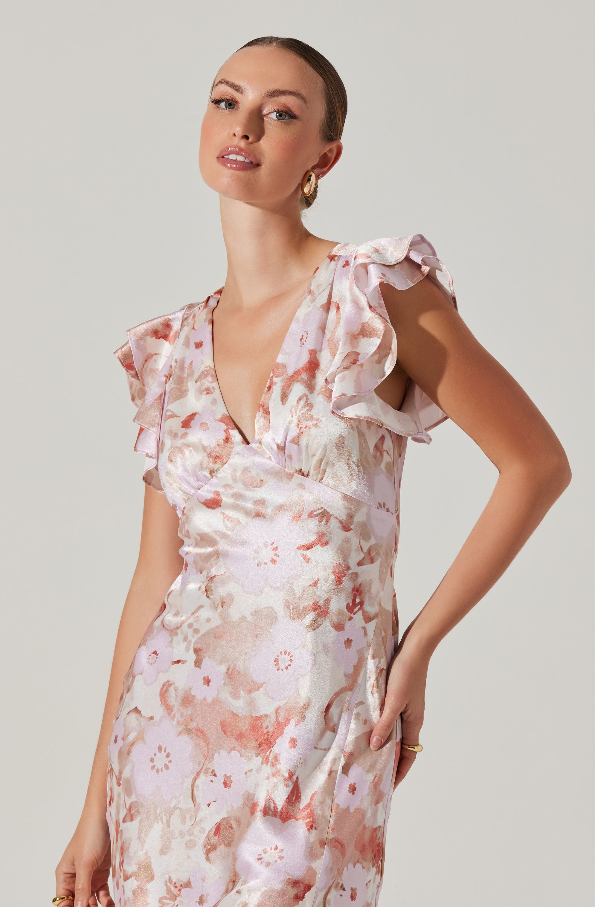 A woman is wearing a sleeveless dress with a V-neckline. The dress features a soft, watercolor floral pattern in shades of pink, peach, and beige, with ruffled details on the shoulders that add a feminine touch.