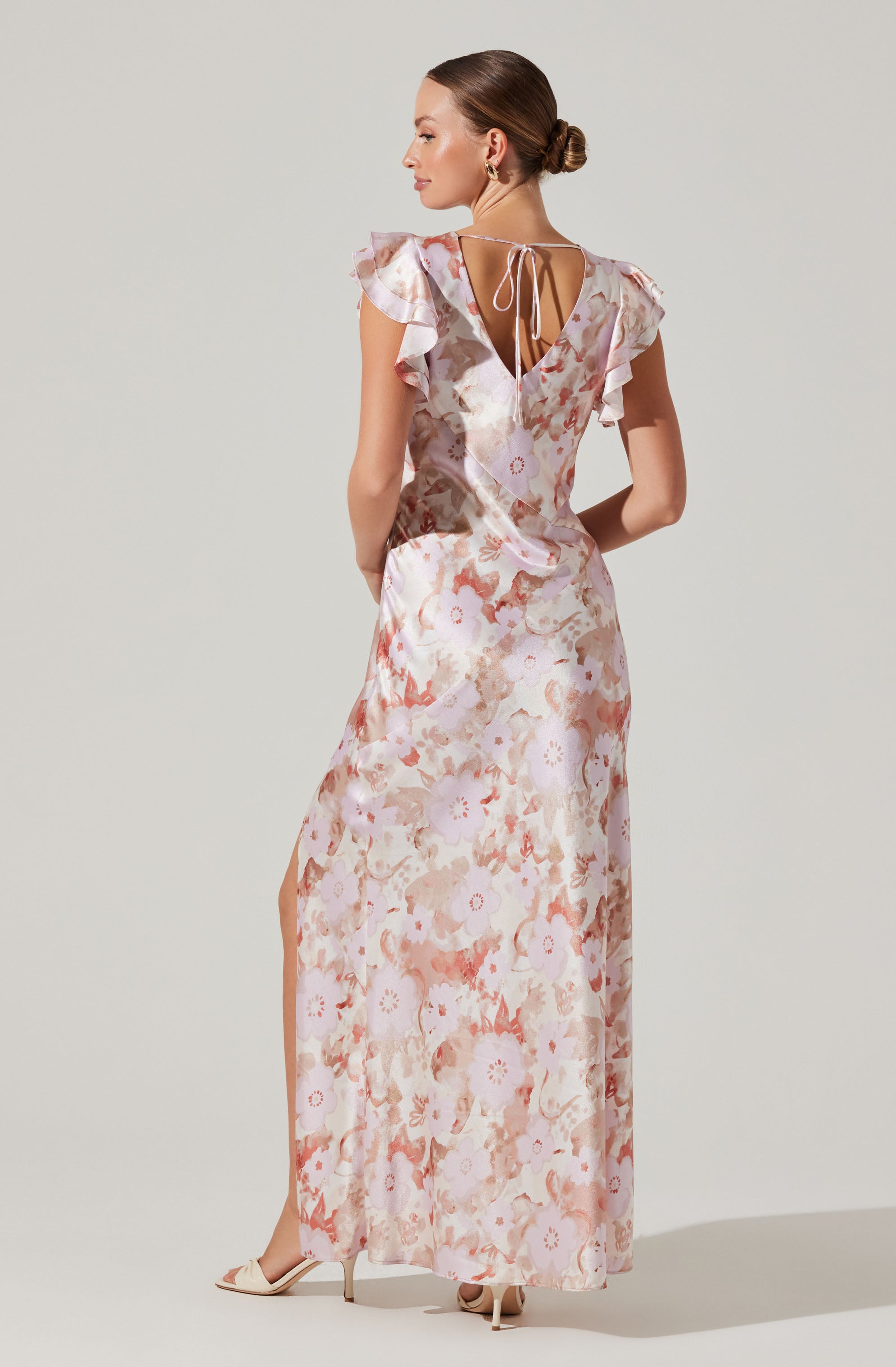 A woman is wearing a full-length, flowing dress with a soft floral pattern in shades of pink, cream, and beige. The dress features ruffled short sleeves and a V-shaped neckline at the back, which ties with a delicate string. The fabric appears lightweight and silky, giving the dress an elegant and graceful look. She is also wearing high-heeled shoes.