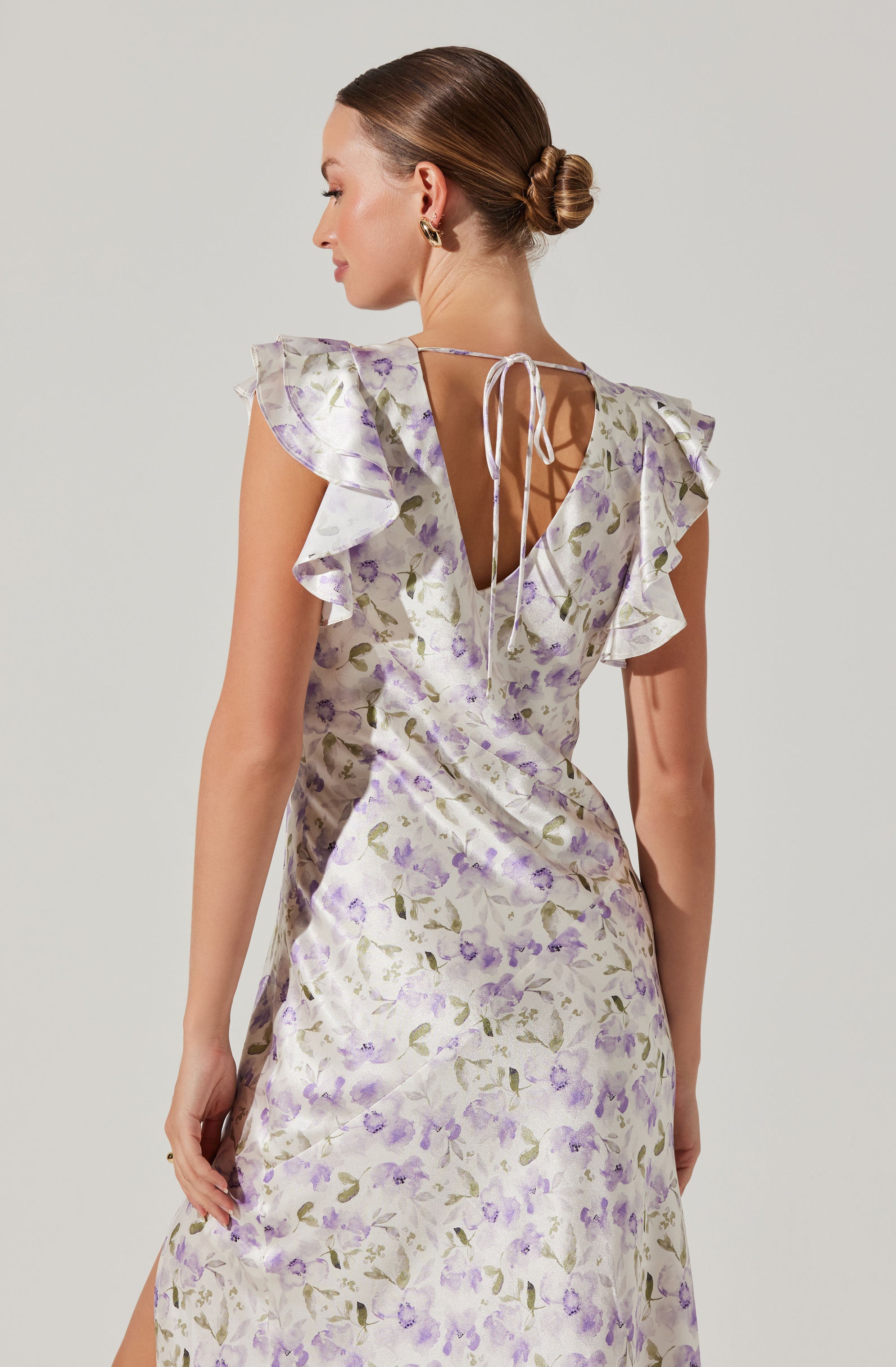 A woman is wearing a sleeveless, knee-length dress with a light-colored background and a floral pattern in shades of purple and green. The dress features ruffled cap sleeves and a V-shaped neckline at the back, which is tied with a delicate ribbon.