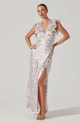 A woman is wearing a long, sleeveless dress with ruffled shoulders and a deep V neckline. The dress features a light, pastel-colored floral pattern with shades of purple and green on a white background. It has a thigh-high slit on the left side, revealing her leg, and she is wearing strappy heeled sandals.
