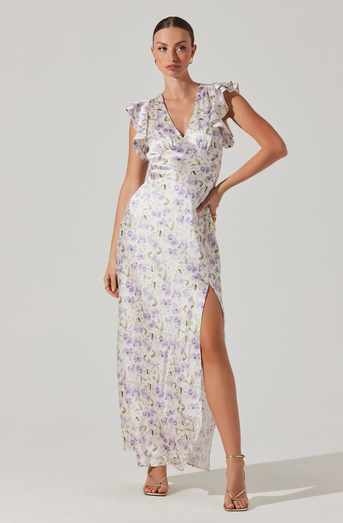 A woman is wearing a long, flowy dress with a delicate floral print in shades of lavender, purple, and green on a light background. The dress features a V-neckline, ruffled short sleeves, and a high slit on one side that extends to the floor. She is also wearing nude-colored high-heeled sandals.