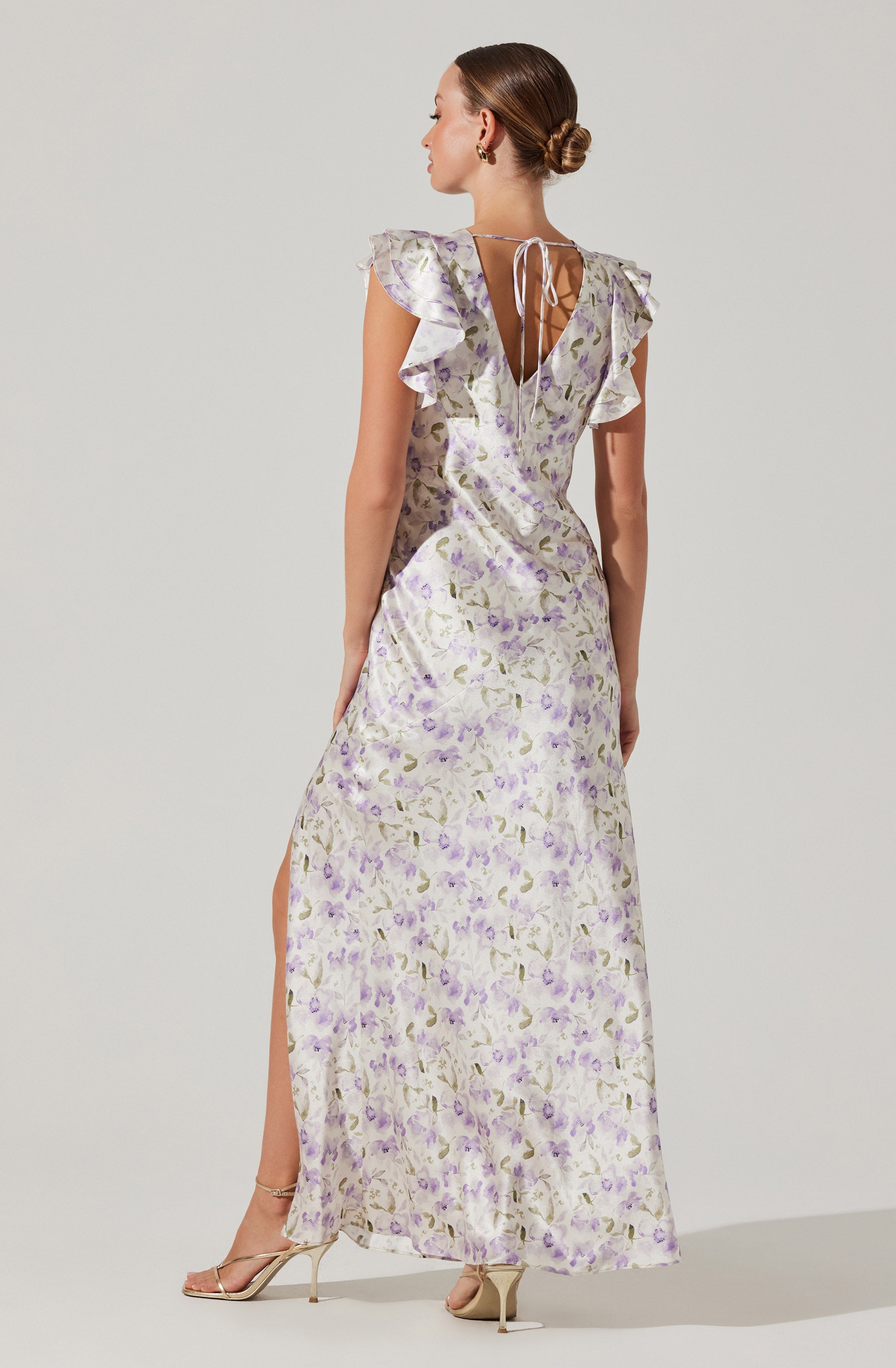 A woman is wearing a long, flowing dress with a light background and a floral pattern featuring shades of purple and green. The dress has ruffled cap sleeves, a V-shaped open back with a thin tie at the top, and a slit on the side. She is also wearing high-heeled sandals.
