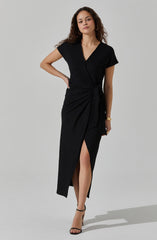 A woman is wearing a black dress with short sleeves and a V-neckline. The dress features a tied waist with a draped detail on one side, creating a slit that reveals her leg. She is also wearing black high-heeled sandals and gold jewelry, including earrings and stacked bracelets.
