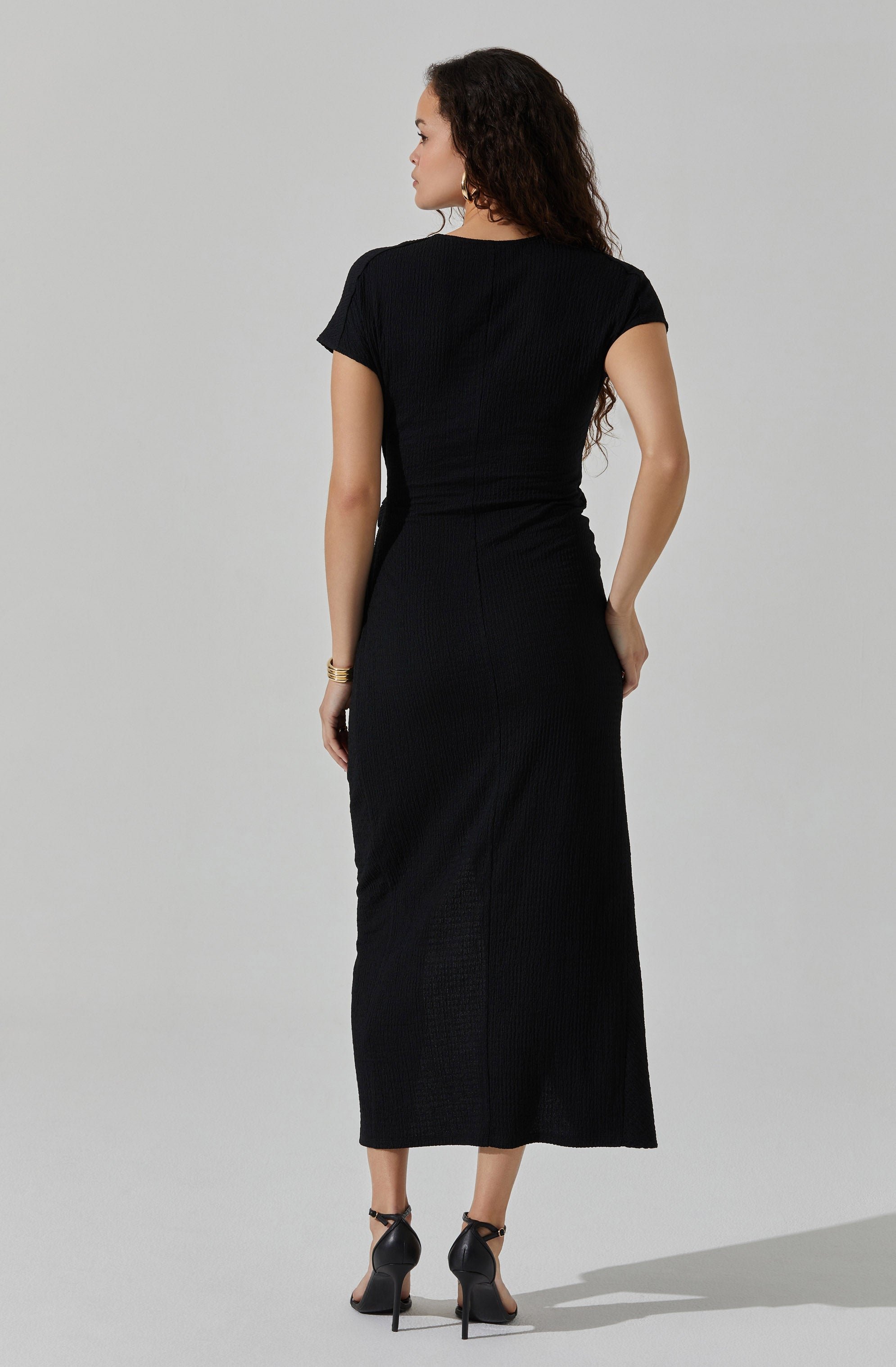 A woman is wearing a fitted black short-sleeve top and a long, black, ribbed skirt that reaches her ankles. She has paired the outfit with high-heeled black shoes and gold jewelry, including bracelets. The overall look is elegant and monochromatic.
