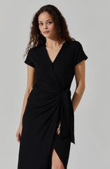 A woman is wearing a black wrap dress with a V-neckline and short sleeves. The dress features a tied detail at the waist and a slit up the front. The fabric has a textured pattern.
