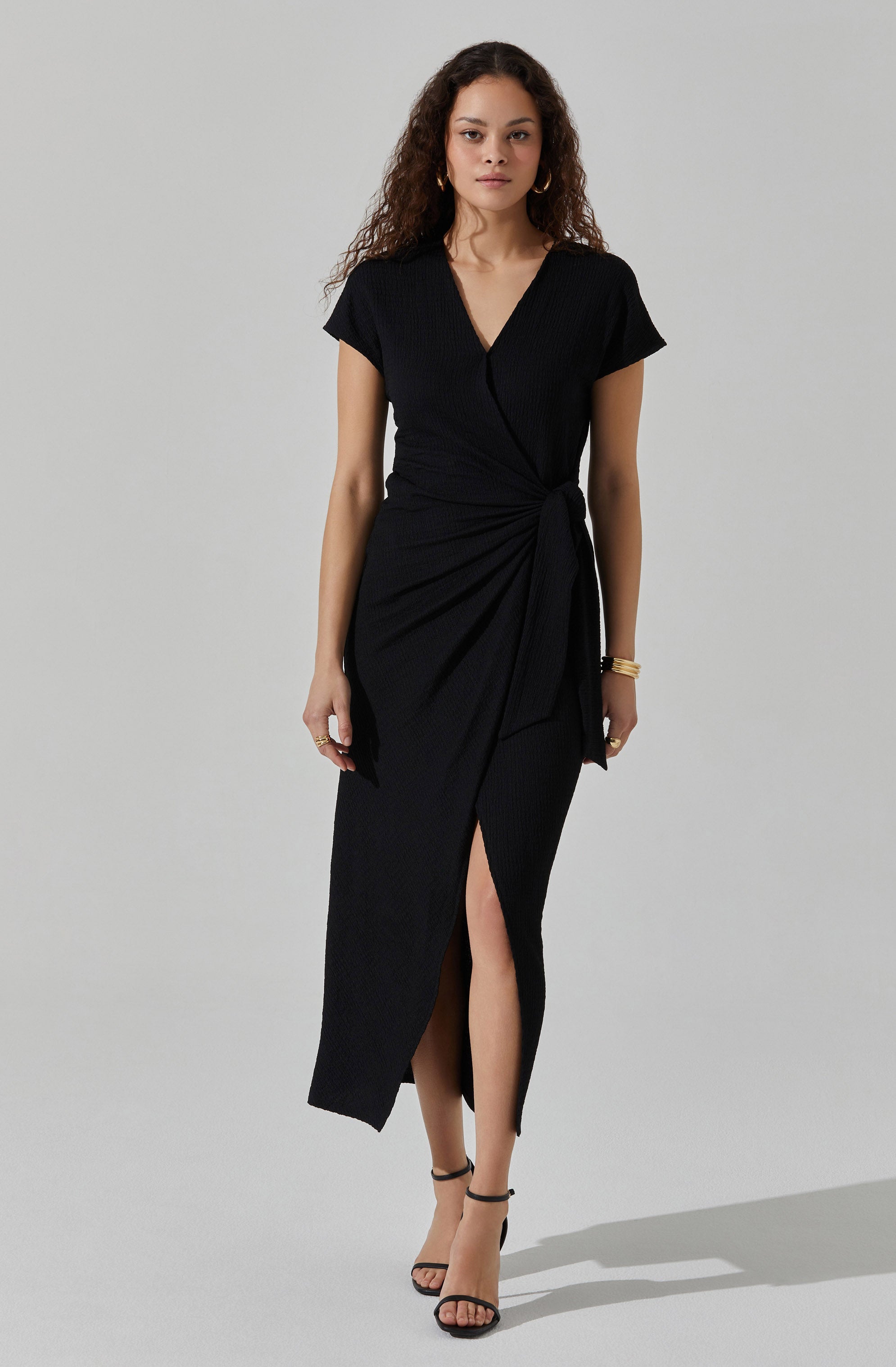 A woman is wearing a black dress with short sleeves and a V-neckline. The dress has a wrap-style design with a tied knot at the waist and a knee-high slit in the front. She is also wearing black strappy high-heeled sandals and gold jewelry, including hoop earrings and bracelets.