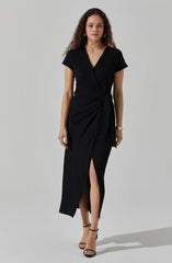 A woman is wearing a black dress with short sleeves and a V-neckline. The dress has a wrap-style design with a tied knot at the waist and a knee-high slit in the front. She is also wearing black strappy high-heeled sandals and gold jewelry, including hoop earrings and bracelets.