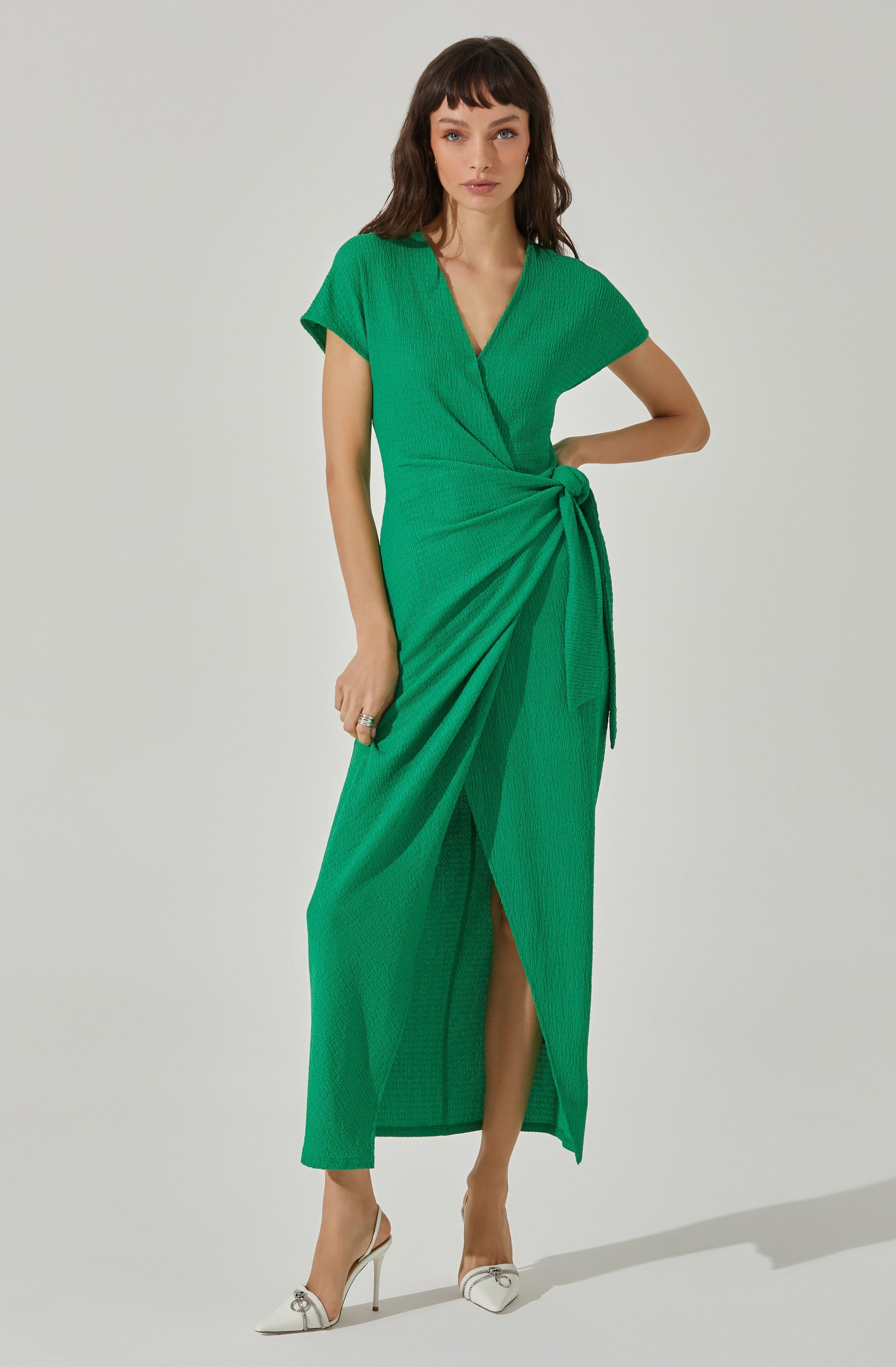 A woman is wearing a vibrant green dress with a wrap-style design. The dress features short sleeves, a V-neckline, and a tied waist that creates a gathered effect, with the fabric draping elegantly to the hem. The skirt has a slit that reveals her legs. She is wearing white high-heeled shoes with pointed toes and decorative embellishments.