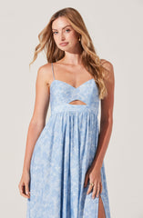 The dress features a light blue, floral patterned fabric with thin spaghetti straps. The bodice has a sweetheart neckline with a small cutout detail just below the bust. The waistline is gathered, creating a flowing, gathered skirt with a slit on one side.