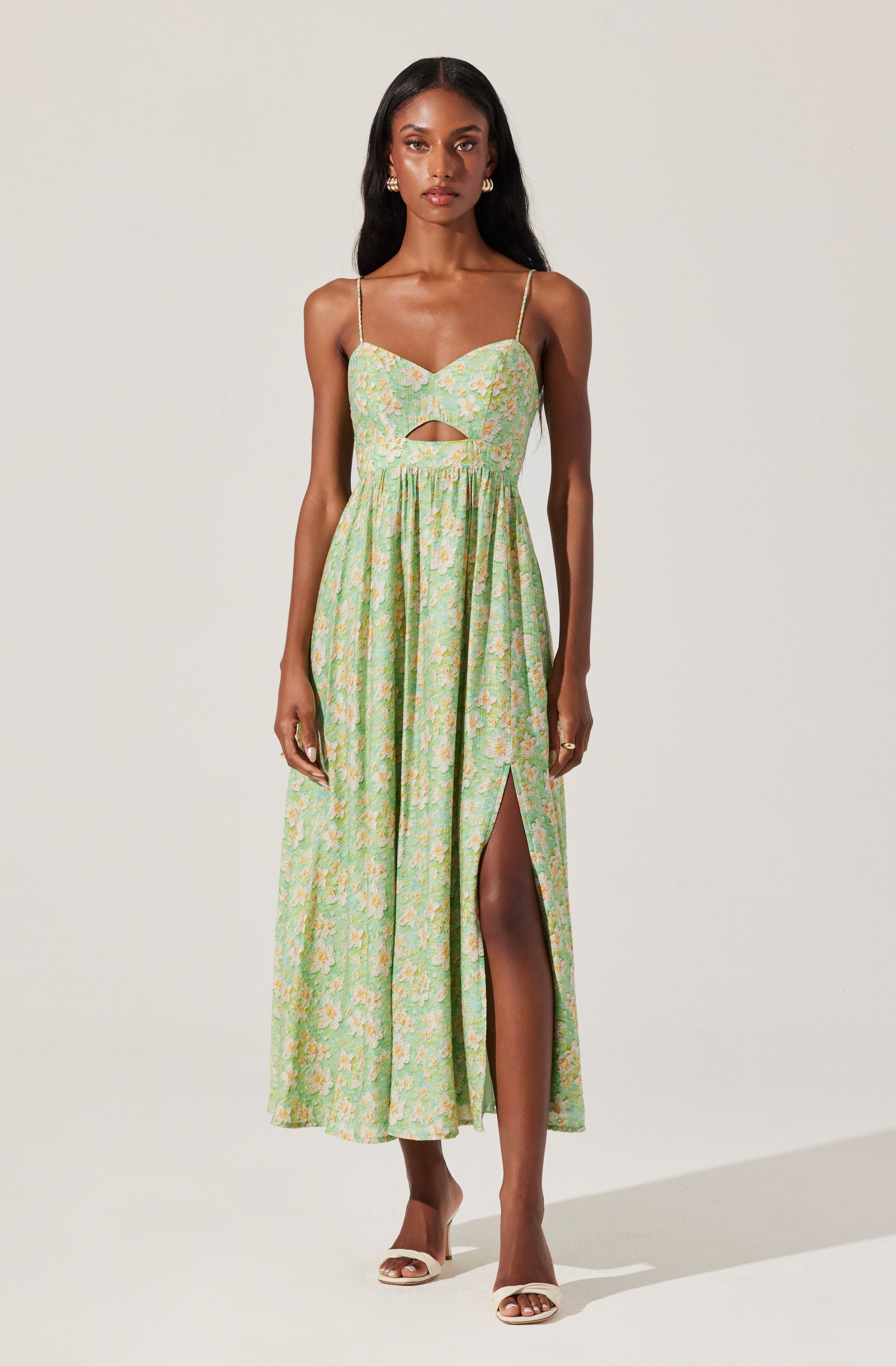 A woman is wearing a sleeveless, ankle-length dress with thin spaghetti straps. The dress features a fitted bodice with a small keyhole cutout in the center. The fabric has a light, pastel-colored floral pattern, primarily in shades of green, with hints of yellow and pink. The skirt is flowing, with a high slit on one side revealing her leg. She completes the look with open-toed, white heeled sandals and gold earrings.