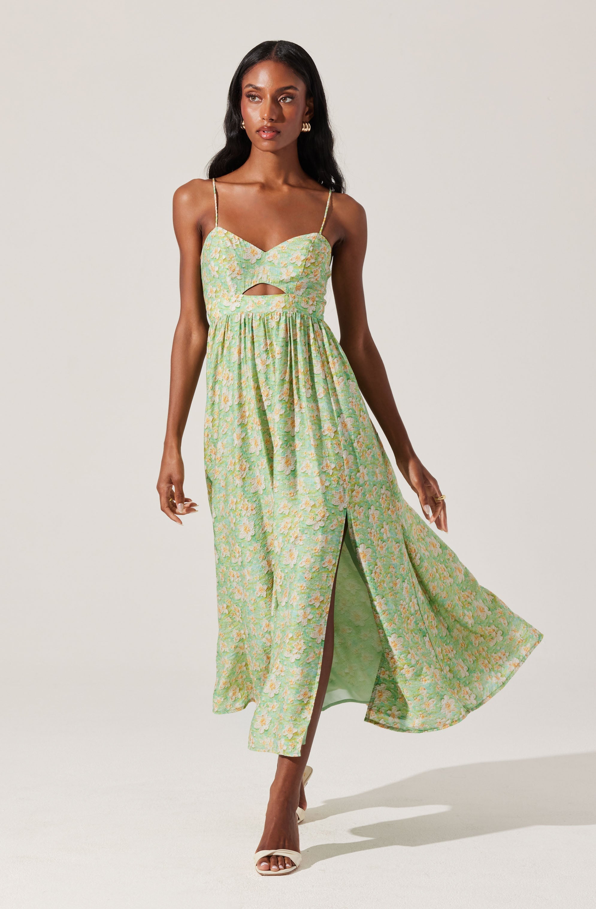 A woman is wearing a sleeveless, ankle-length dress with thin spaghetti straps. The dress features a fitted bodice with a slight sweetheart neckline, a small cutout below the bust, and a high slit at the front revealing one leg. The fabric has a light pastel floral pattern in shades of green, pink, yellow, and white.