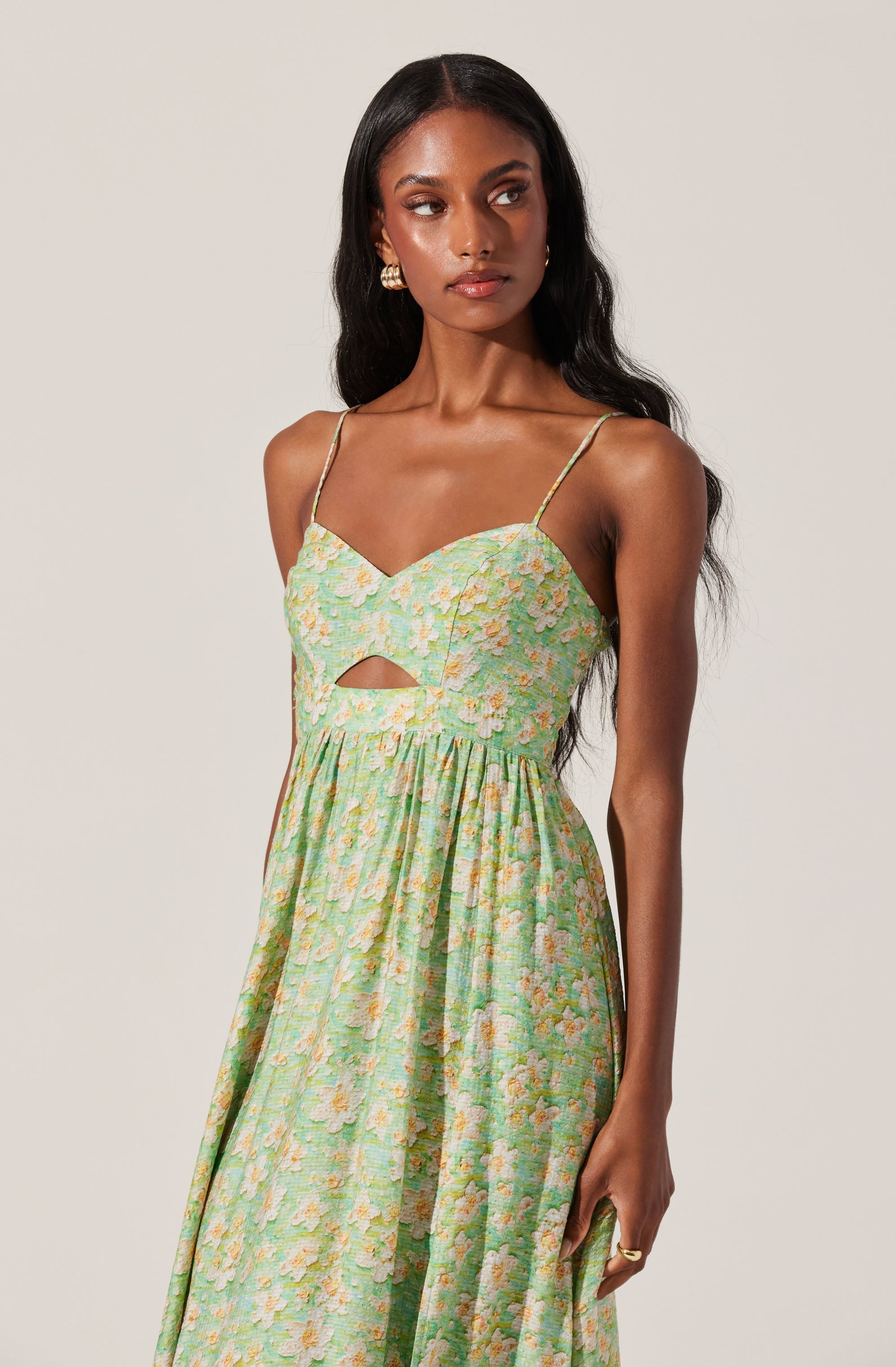 A woman is wearing a sleeveless, ankle-length dress with thin spaghetti straps. The dress features a pastel green background with a floral pattern in soft pink, peach, and cream colors. It has a cut-out detail just below the bust. The fabric appears lightweight and flowy.