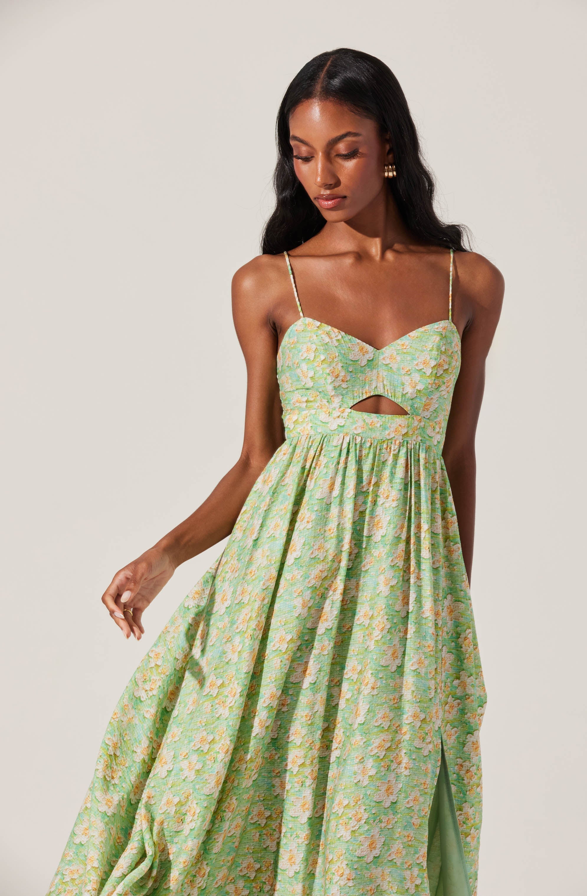 A woman is wearing a sleeveless dress with thin spaghetti straps. The dress features a sweetheart neckline and a small cut-out detail just below the bust. The fabric of the dress has a light, pastel floral pattern in shades of green, yellow, and pink. The skirt is long, flowing, and gathered at the waist, creating a full, elegant silhouette.