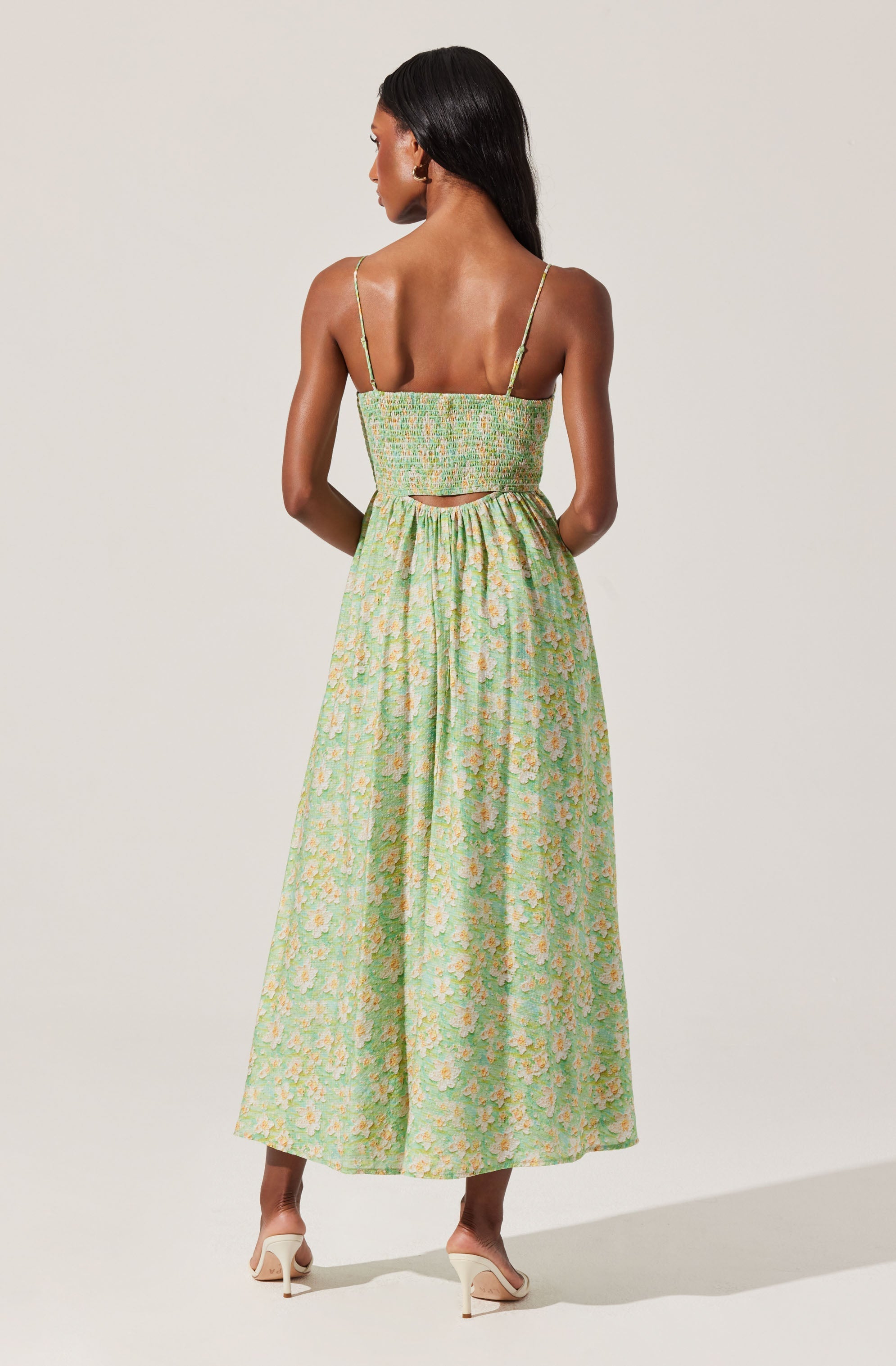 A woman is wearing a sleeveless, green dress with a floral pattern, featuring thin spaghetti straps, a fitted bodice, a small cutout at the back, and a long, flowing skirt that reaches her ankles. She is also wearing beige high-heeled shoes.