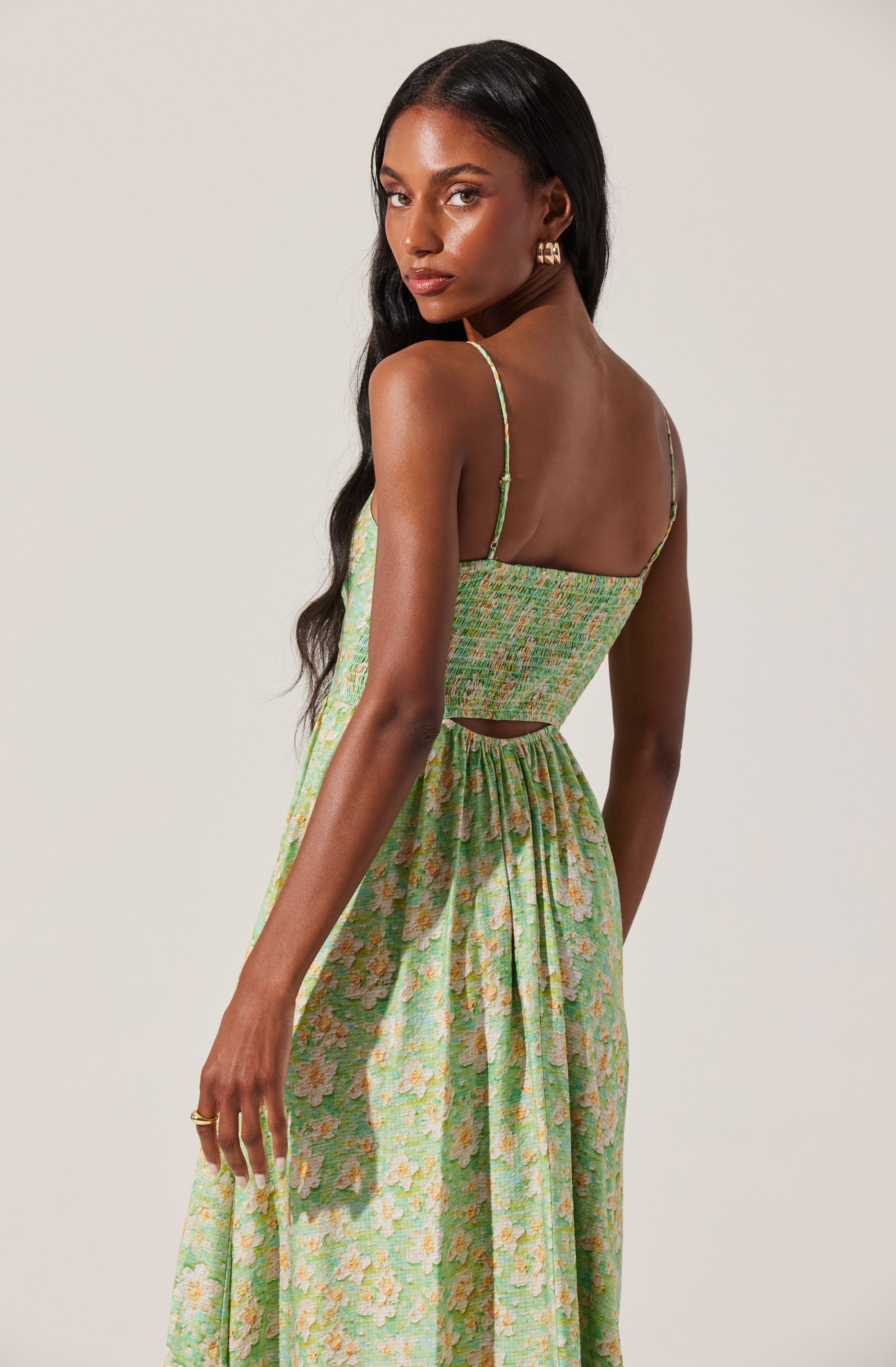 A woman is wearing a sleeveless, spaghetti-strap top and a matching long skirt, both made from a light, pastel-colored fabric featuring a floral pattern with shades of green, pink, and yellow. The top is fitted and cropped with a smocked design, and the skirt has a gathered waistline creating a flowing silhouette.