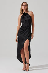A woman is wearing a sleeveless black dress with a halter neckline adorned with a large fabric flower. The dress features a keyhole opening at the neck and a high slit on the left side, revealing her leg. She is accessorized with gold bracelets and black high-heeled sandals.