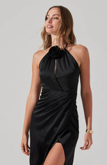 A woman is wearing a black satin evening gown with a halter neckline featuring a large fabric flower detail at the neck. The dress has a keyhole opening at the chest and a side slit that reveals her leg. The fabric drapes elegantly, adding to the dress's sophisticated look.