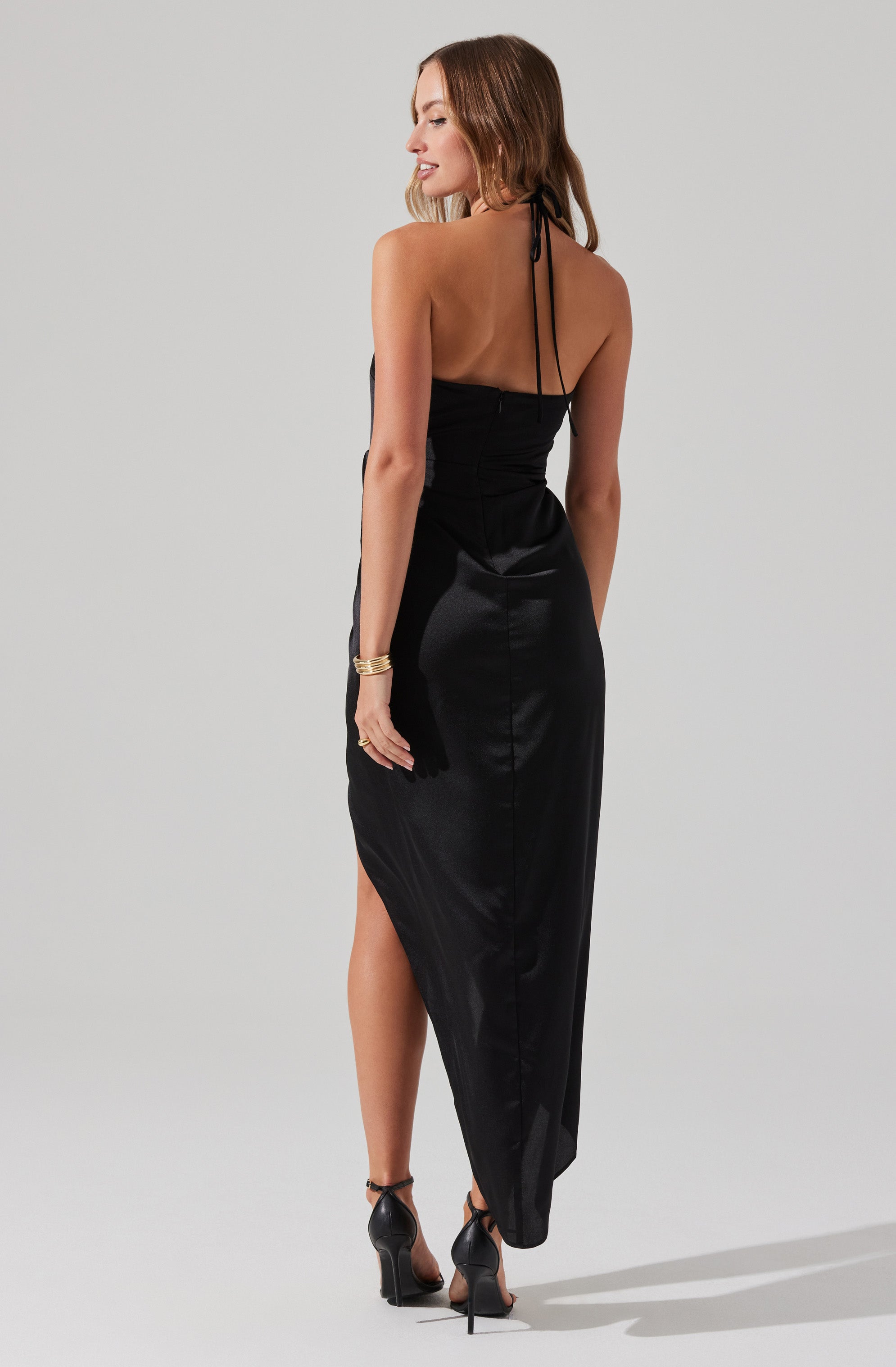 A woman is wearing a long, black satin dress with a fitted bodice and a halter neckline that ties around her neck. The dress has a high slit on one side, revealing her leg. She is also wearing black high-heeled sandals and gold bracelets on her left wrist.