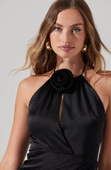 A woman is wearing a sleeveless black satin dress with a halter neckline, featuring a black fabric flower detail at the neck. The dress has a keyhole opening in the front. She accessorizes with large gold hoop earrings.