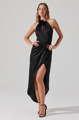 A woman is wearing a floor-length black dress with a halter neckline that features a large fabric flower at the center. The dress has a keyhole opening at the chest and a high slit on one side, revealing her leg. She pairs the outfit with black high-heeled sandals with ankle straps and wears a gold bracelet on her wrist.