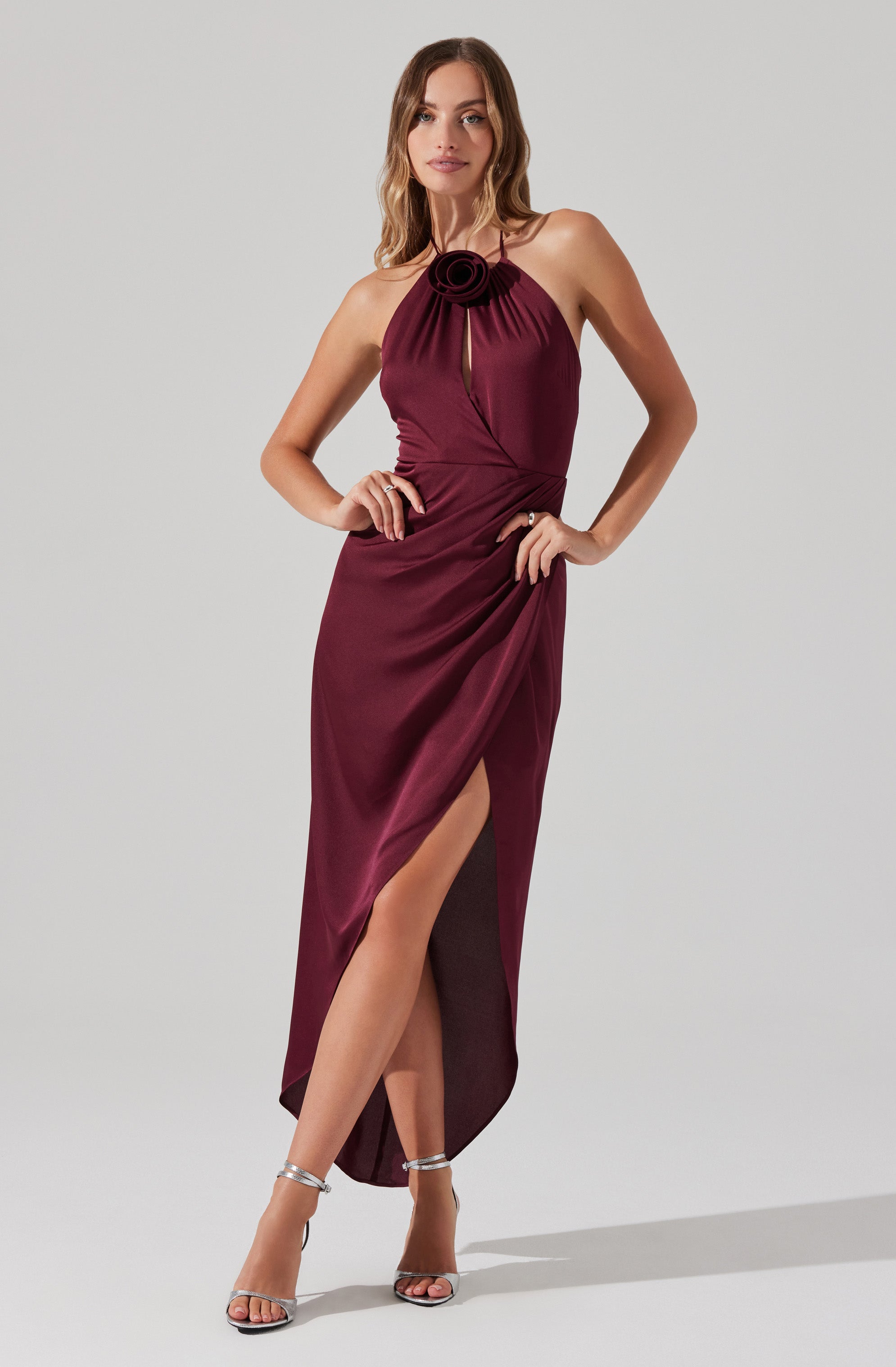 A woman is wearing a sleeveless, floor-length burgundy gown with a halter neckline. The bodice features a fabric flower detail at the center of the neckline, and the dress has a slit up the front, revealing her legs and silver high-heeled sandals.