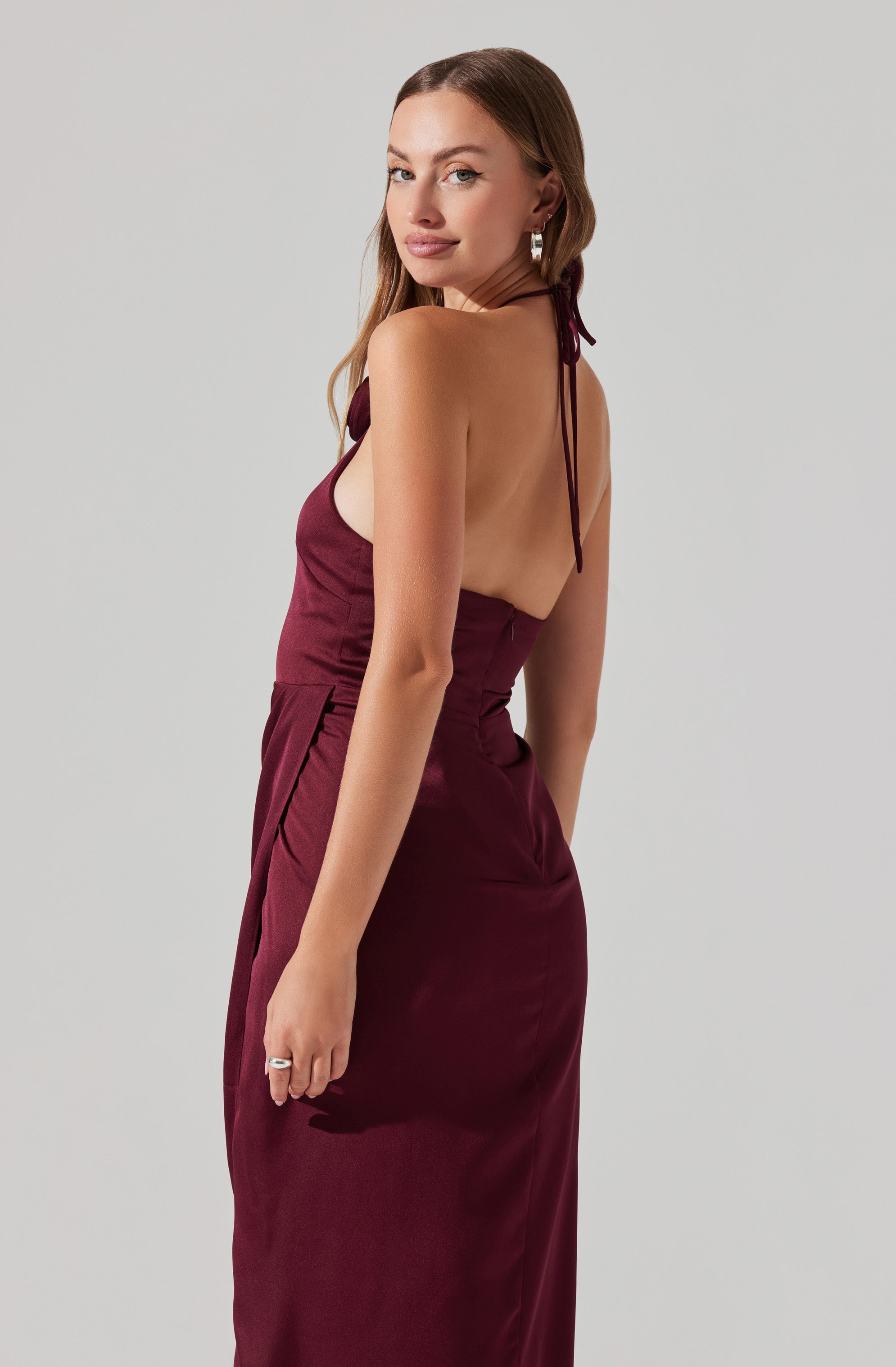 A woman is wearing a sleeveless, floor-length dress in a deep burgundy color. The dress features thin shoulder straps that tie at the back of her neck, leaving her shoulders and back mostly exposed.