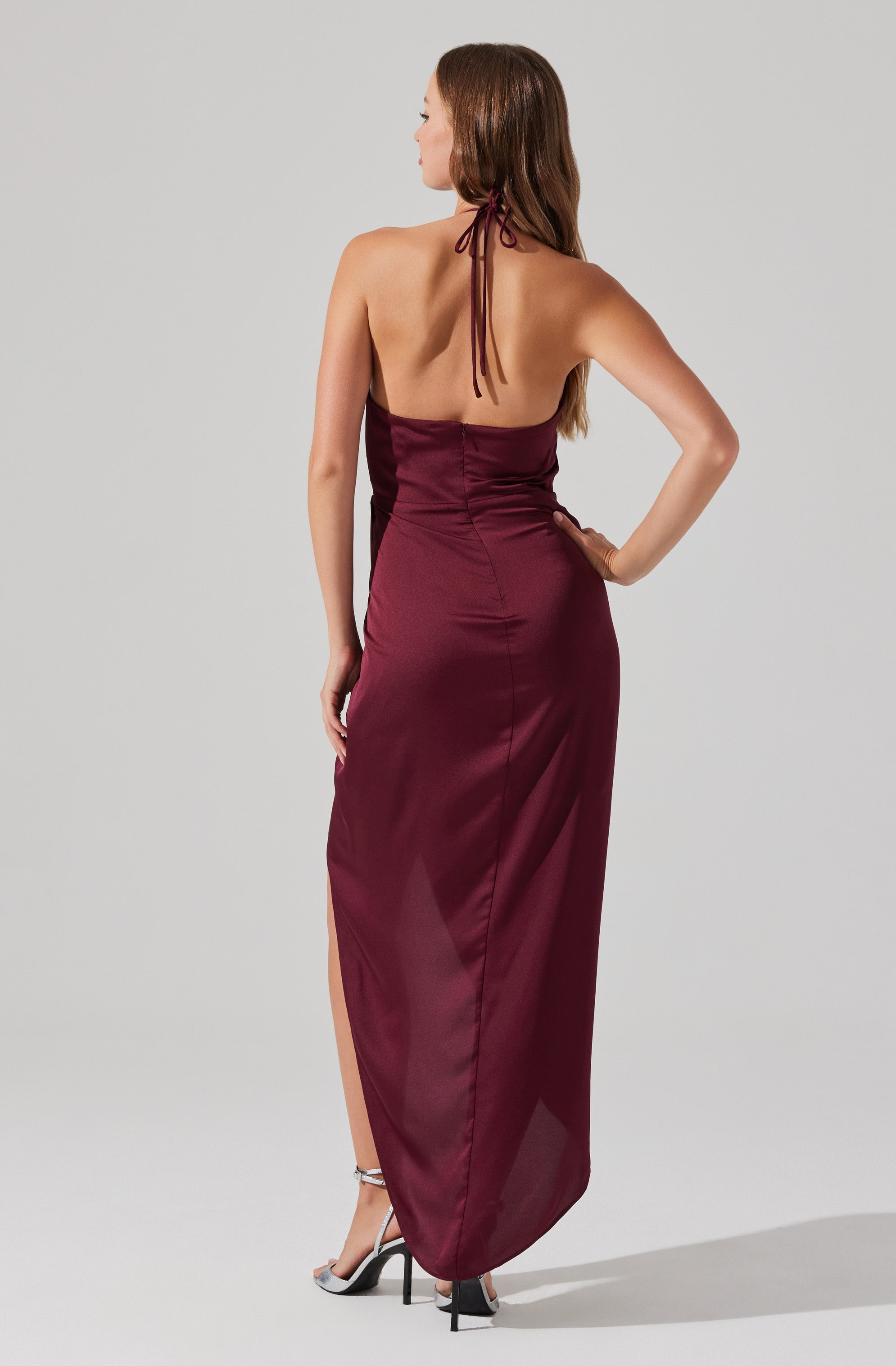 A woman is wearing a sleeveless, halter-neck dress in a deep burgundy color. The dress features a fitted bodice with ruched detailing and a long flowing skirt with a high slit on one side. She is standing with one hand on her hip, and her hair is styled long and loose.