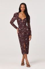 A woman is wearing a long, fitted dress with a dark brown base color and a floral pattern in shades of light purple and gray. The dress features long sleeves with puffed shoulders, a sweetheart neckline with a small keyhole cutout in the center, and gathers along the torso for a ruched effect. It also has a slit at the front of the skirt, reaching to mid-calf. She is wearing strappy black high-heeled sandals.