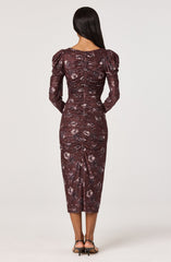 A woman is wearing a fitted, long-sleeved dress with a dark maroon base color and a floral pattern in shades of pink and white. The dress features ruched detailing along the center back and down the length of the skirt, with puffed shoulders adding volume to the sleeves. She is also wearing black open-toe heels.