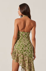 A woman is wearing a sleeveless dress with a halter neckline. The dress features a floral pattern in shades of green and light pink, with an asymmetrical ruffled hem. The back of the dress is open, revealing her bare shoulders and upper back.