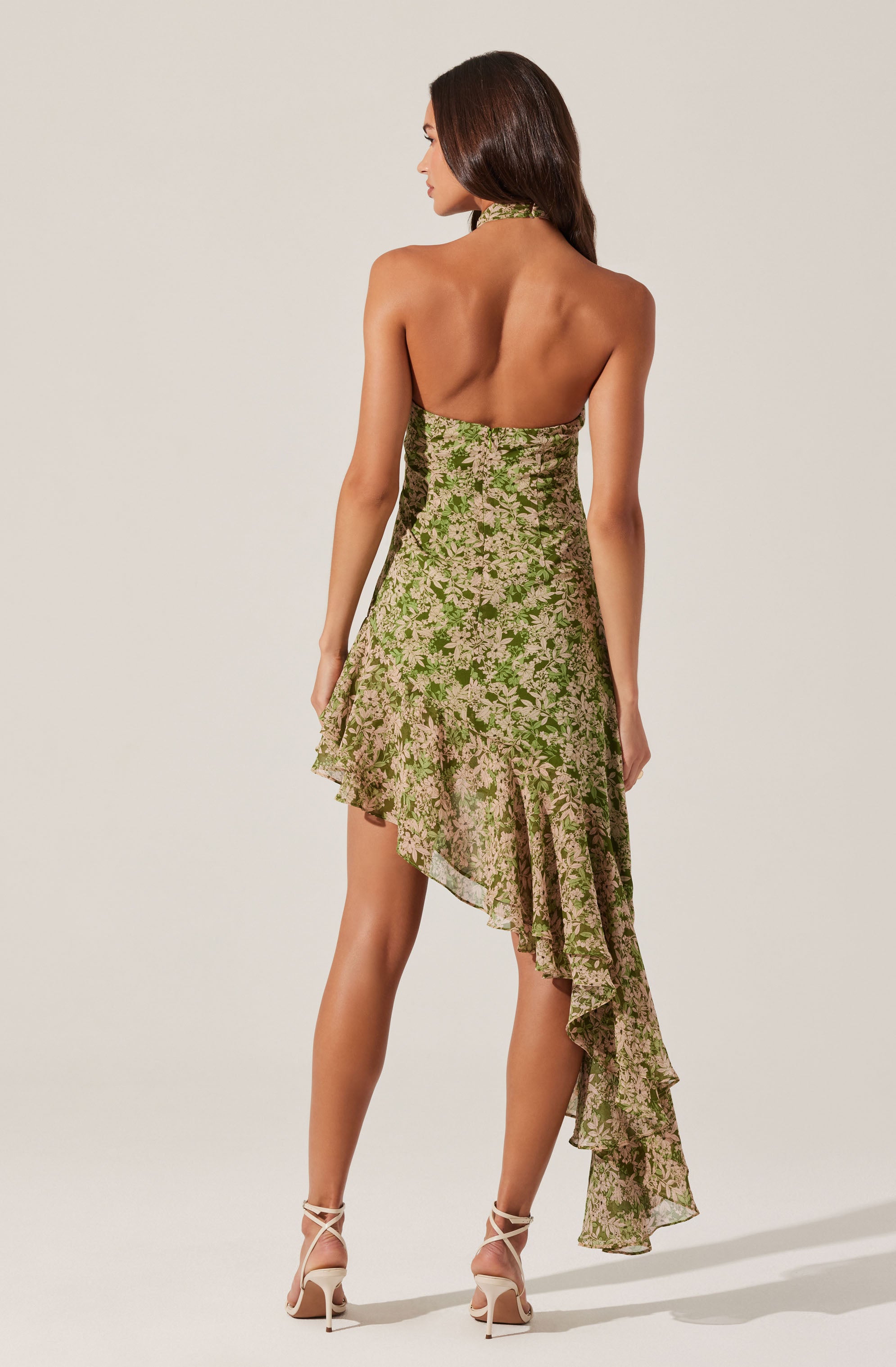 The dress is a strapless, asymmetrical, floral-printed piece featuring green and beige colors. It has a fitted bodice and a ruffled, layered skirt that is shorter in the front and longer in the back, creating a high-low hemline. The floral pattern is dense with small flowers, and the fabric appears lightweight and flowy. The model is wearing nude, strappy high-heeled sandals.