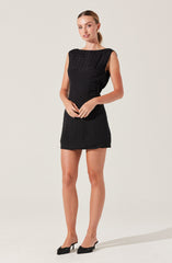 A woman is wearing a sleeveless black dress with a textured pattern on the upper part. The dress is fitted and short, ending above her knees. She pairs it with black high-heeled shoes that have a bow detail on the front.