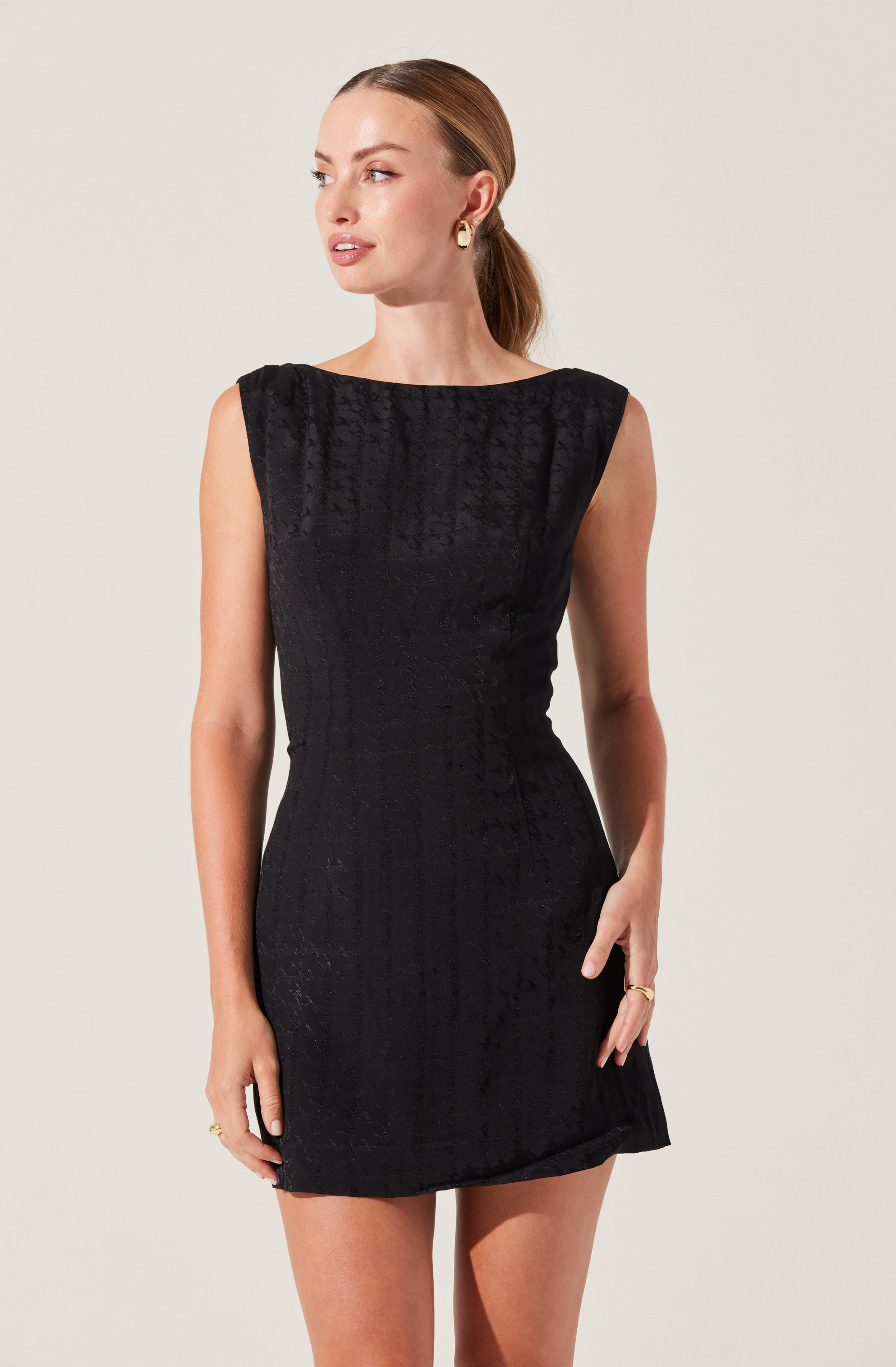 A woman is wearing a sleeveless, black dress with a subtle textured pattern. The dress has a round neckline and a fitted silhouette that falls above the knees. She accessorizes with gold hoop earrings and rings on her fingers.