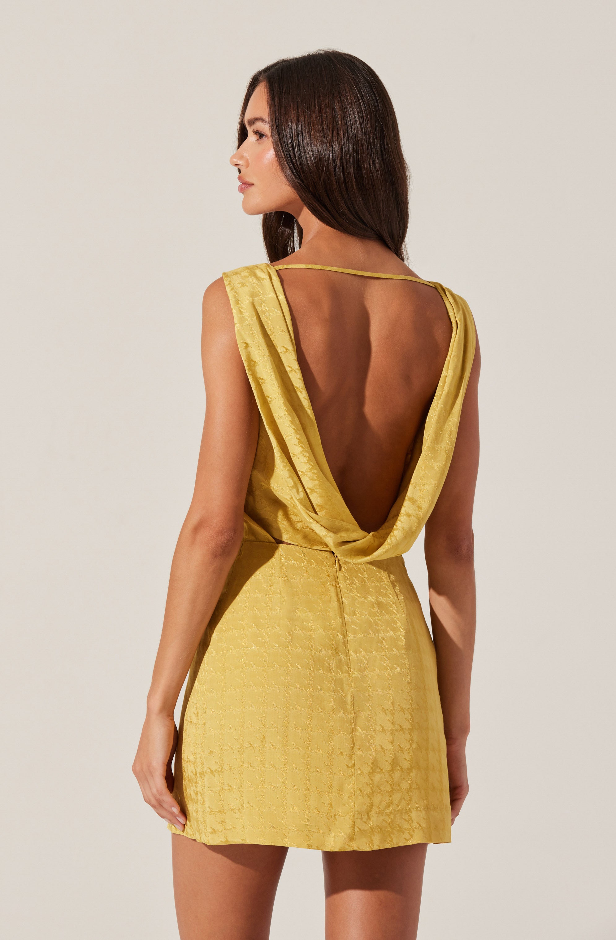 A woman is wearing a sleeveless yellow dress made of shiny, textured fabric. The dress features a deep, open back with draped fabric around the shoulders and an adjustable strap across the top of the open back. The dress is fitted at the waist and flares out slightly at the hem.