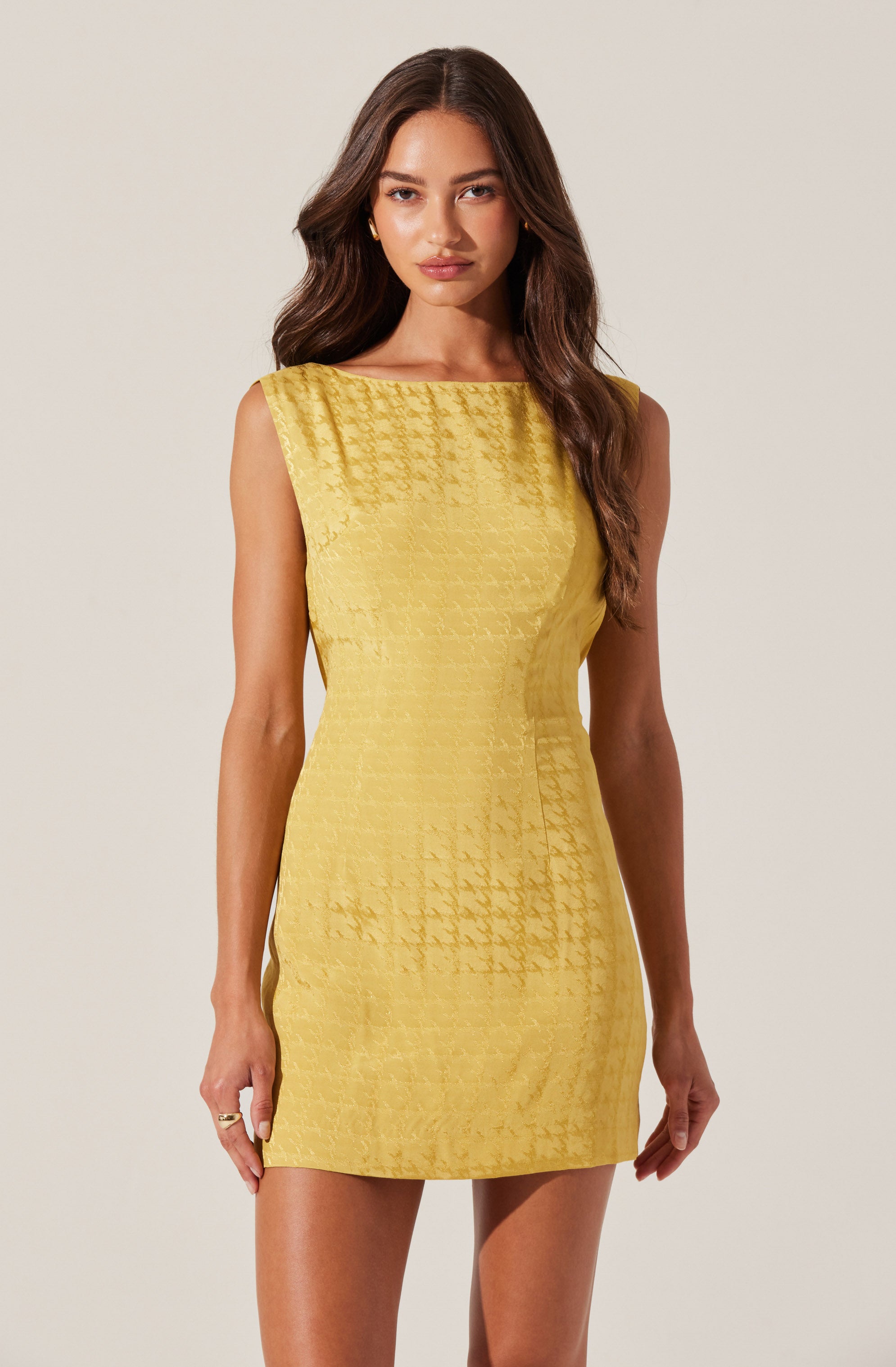 A woman is wearing a sleeveless, fitted yellow dress with a textured, checkered pattern. The dress has a boat neckline and falls above the knees. She has long, wavy brown hair and is standing against a plain, light-colored background.