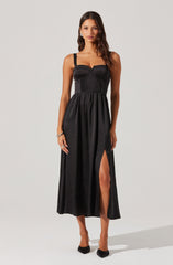 A woman is wearing a black sleeveless dress with wide shoulder straps. The bodice features vertical pinstripes and a fitted, structured design that accentuates the waist. The skirt of the dress is long and flowing, with a high slit on one side revealing her leg. She is also wearing black pointed-toe heels and a bracelet on her right wrist.
