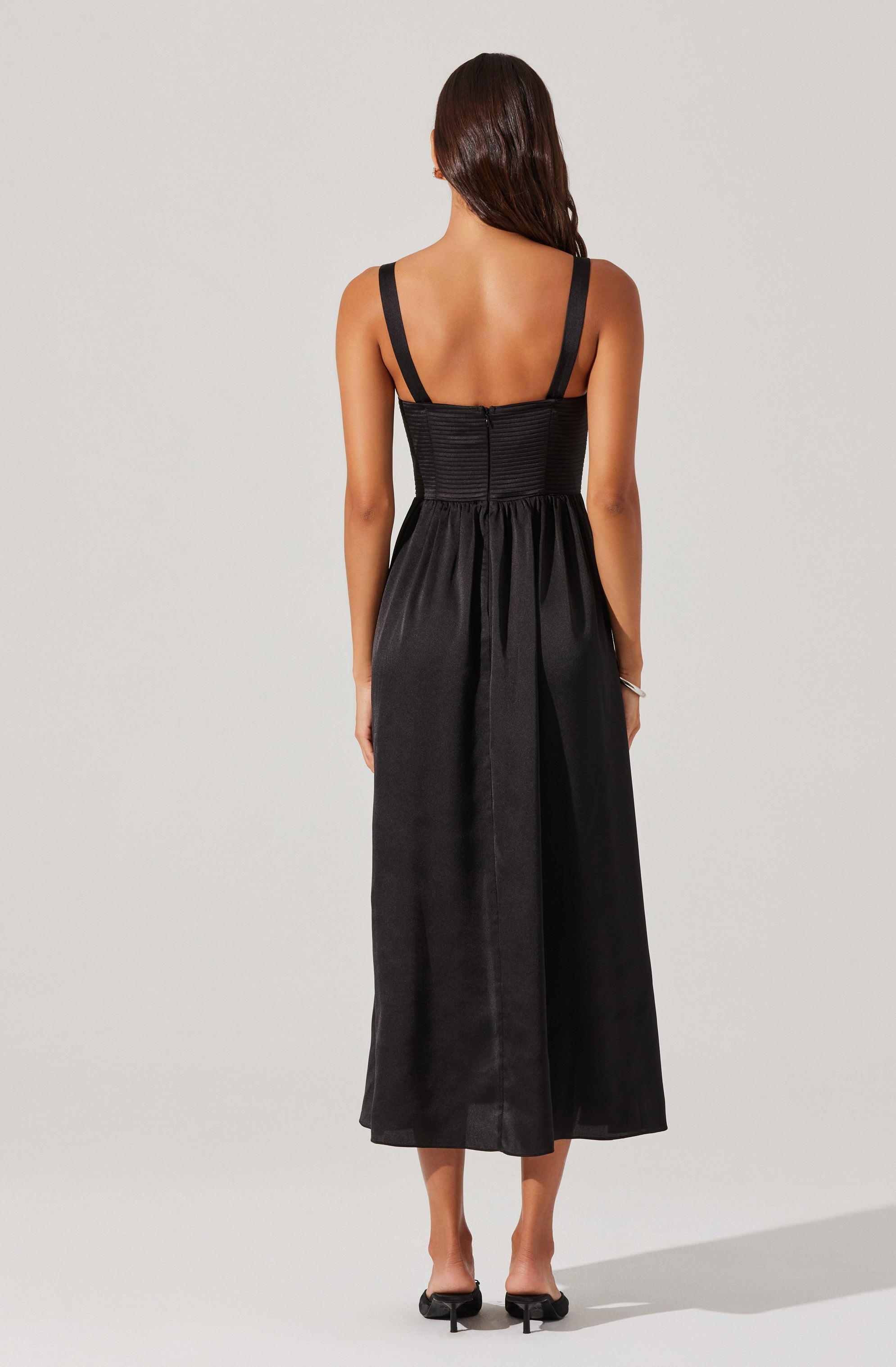 The dress is black with wide shoulder straps and a fitted bodice with horizontal ruching details. The skirt is long and flowy, reaching mid-calf length, made of a shiny, satin-like fabric. The back of the dress features a zipper closure. The overall style is elegant and sophisticated.