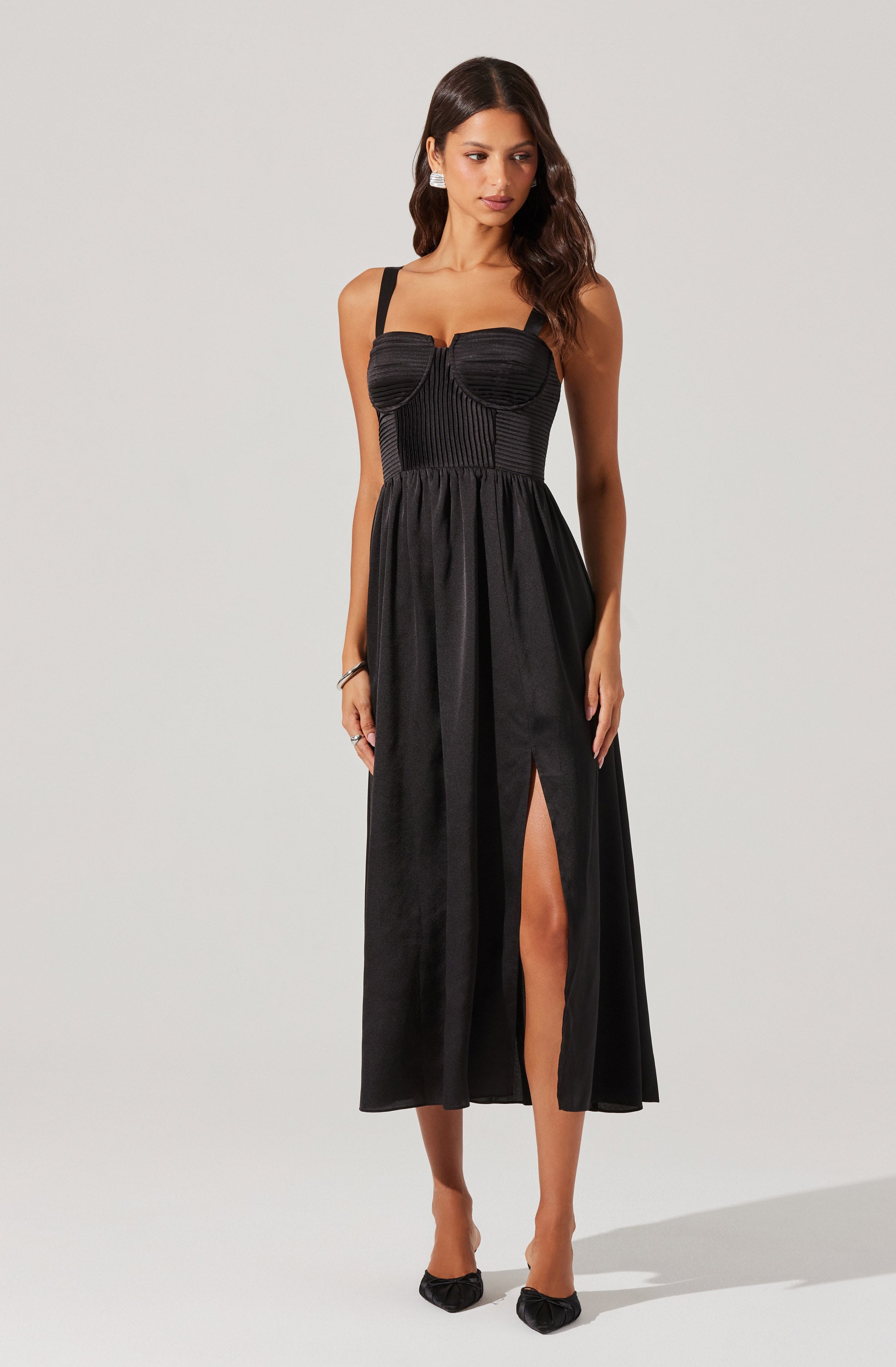 A woman is wearing a black sleeveless dress with wide shoulder straps. The bodice features vertical black stripes and a sweetheart neckline. The dress has a fitted waist, and the skirt flows down to the ankles with a high slit on one side, revealing her leg. She is also wearing black pointed-toe heels.