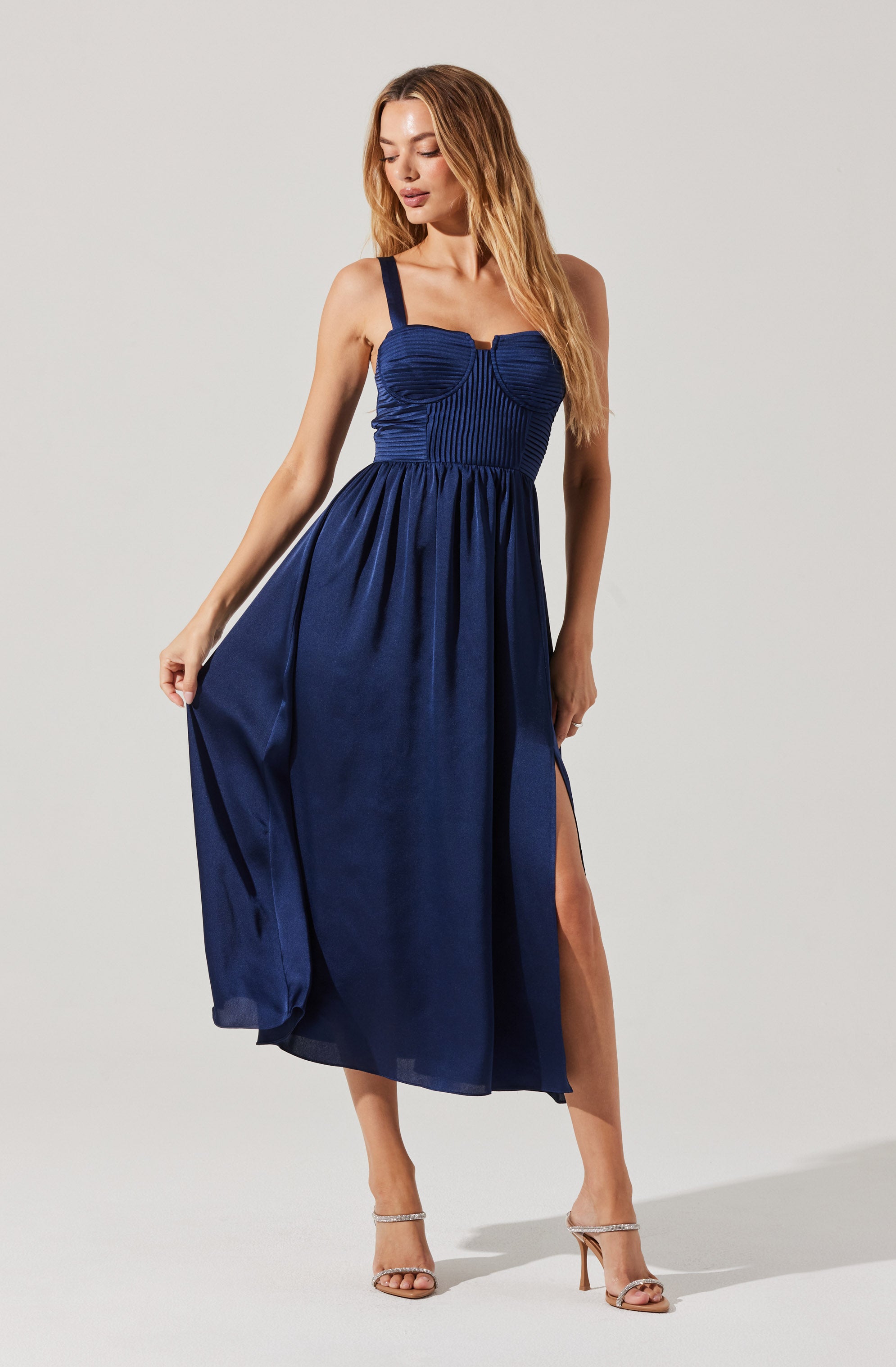 A woman is wearing a sleeveless, deep blue dress with wide shoulder straps. The bodice features vertical and horizontal textured patterns, creating a fitted look. The skirt of the dress is full and reaches down to her ankles, with a slit on the side revealing her leg. She is also wearing strappy high-heeled sandals.