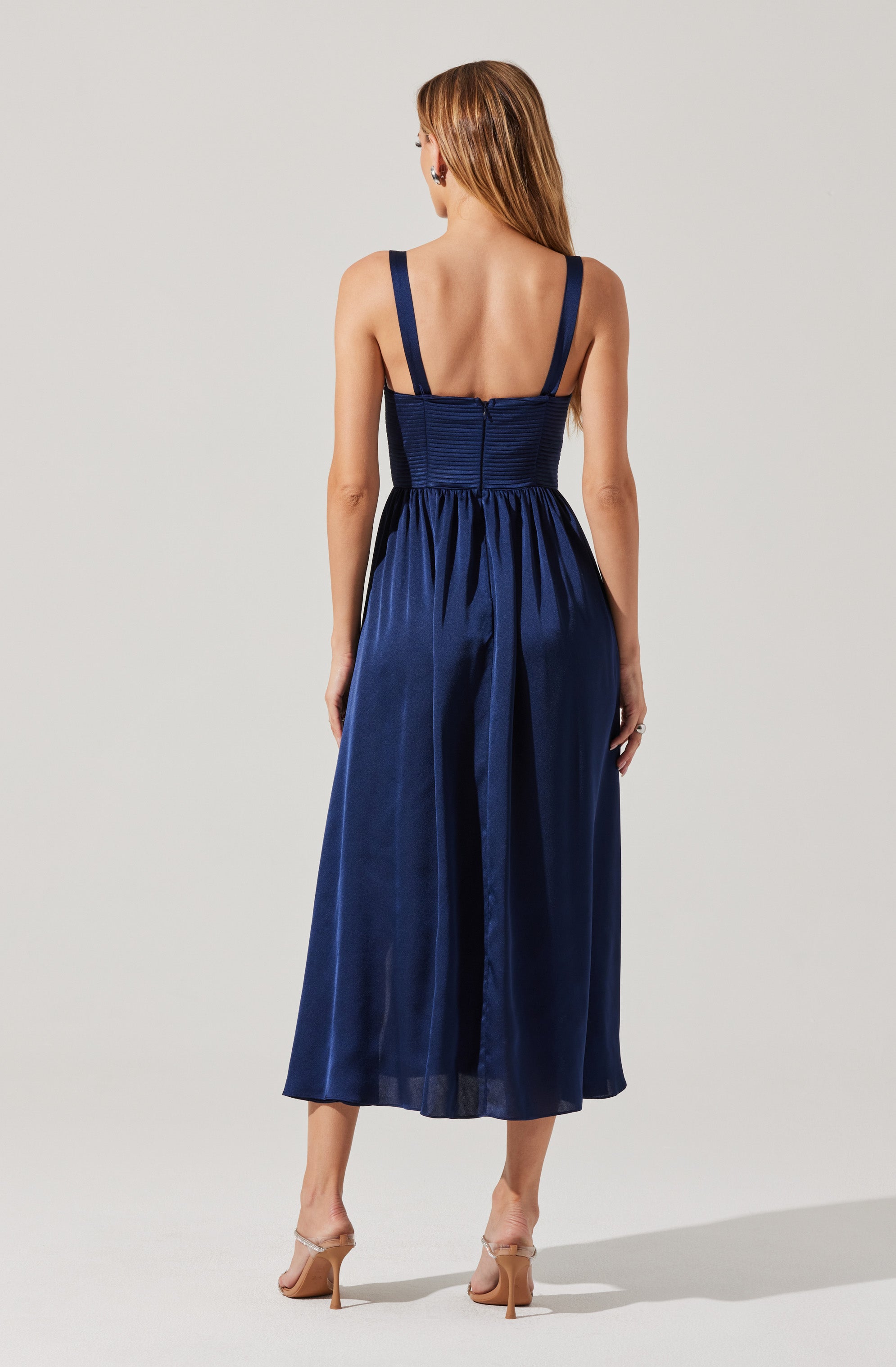 A woman is wearing a sleeveless, navy blue dress with thick shoulder straps. The bodice is fitted with horizontal ruching details, and the skirt is full and flowing with a slight sheen, reaching mid-calf length. She is also wearing nude high-heeled shoes.
