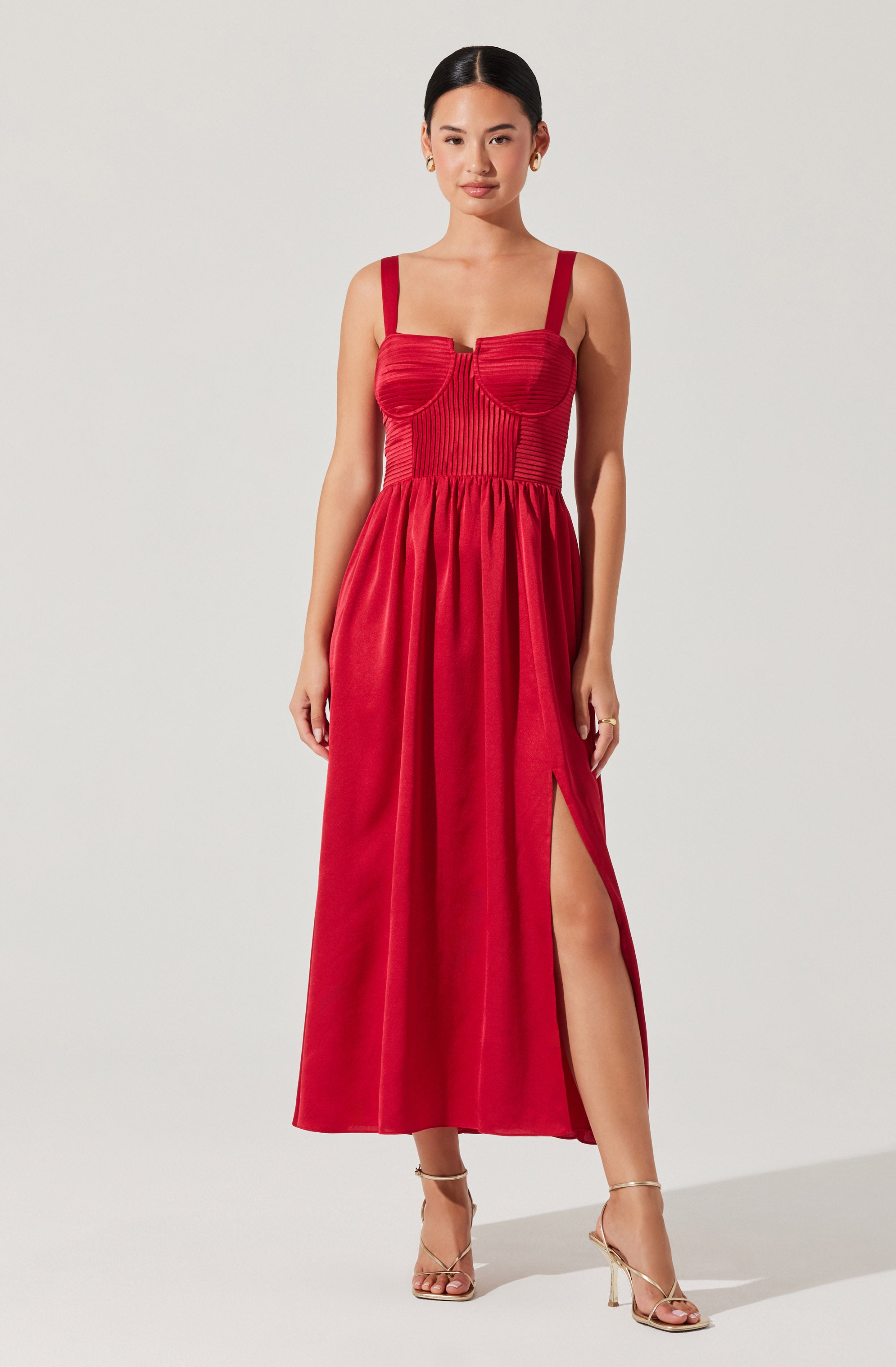 The dress is a sleeveless, floor-length gown in a bold red color. It features a fitted bodice with vertical pleats and structured, curved seam details that enhance the bust area. The dress has wide straps and a high slit on one side, revealing part of the leg. The skirt is gathered at the waist and flows down freely to the floor. The overall style is elegant and sophisticated.
