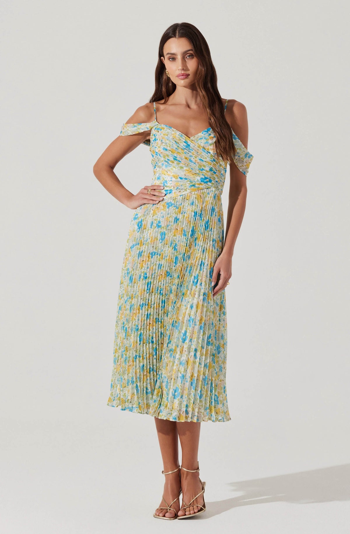 A woman is wearing a sleeveless, ankle-length dress with thin spaghetti straps and off-the-shoulder ruffle sleeves. The dress features a multicolored pattern in shades of yellow, blue, and white, with a fitted waist and a pleated skirt. She is also wearing strappy beige high-heeled sandals.