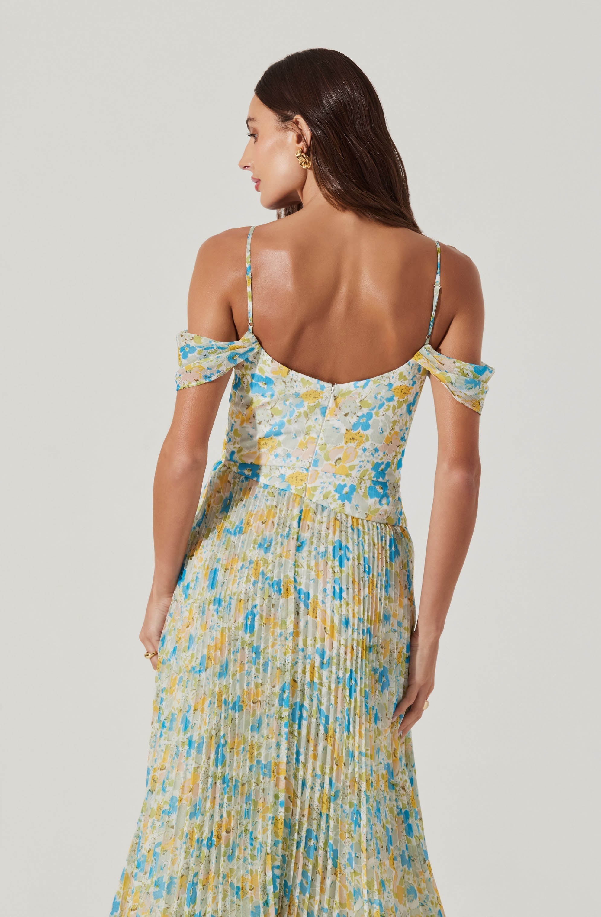 A woman is wearing a sleeveless dress with thin spaghetti straps and off-shoulder bows. The fabric features a multicolored floral pattern with shades of blue, yellow, and white, and the skirt is pleated, creating a textured appearance.