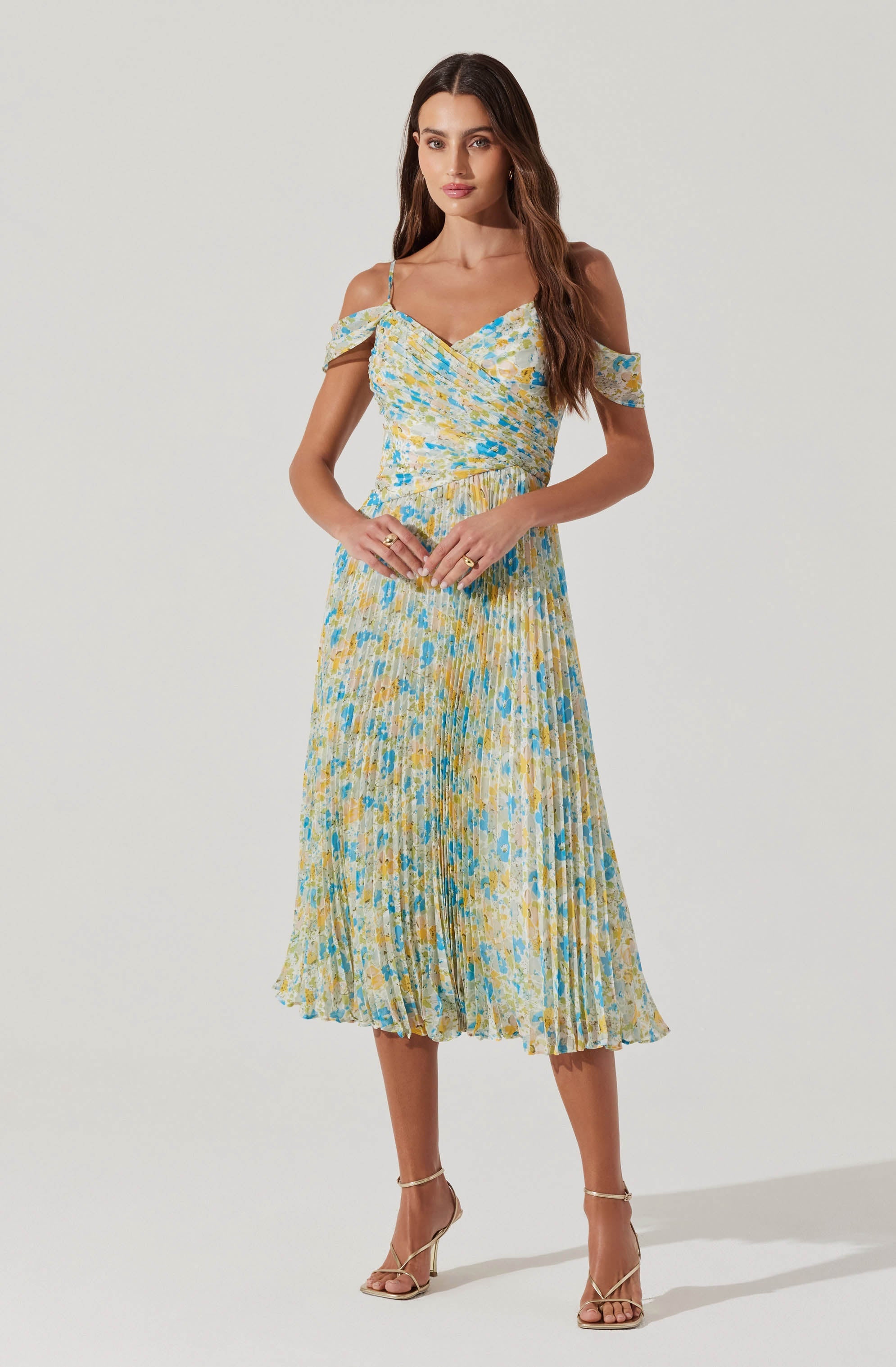 A woman is wearing a light-colored dress with a floral pattern in shades of yellow, blue, and cream. The dress has thin straps, an off-the-shoulder ruffle detail, a fitted waist, and a pleated, midi-length skirt.