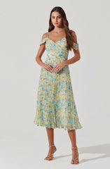 A woman is wearing a light-colored dress with a floral pattern in shades of yellow, blue, and cream. The dress has thin straps, an off-the-shoulder ruffle detail, a fitted waist, and a pleated, midi-length skirt.