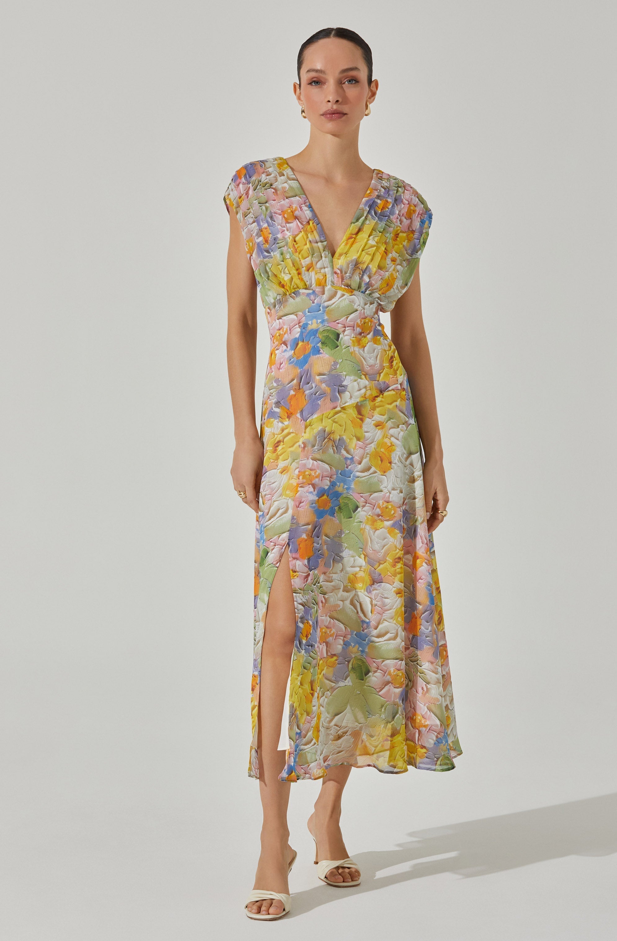 A woman is wearing a sleeveless, long dress with a V-neckline and a floral pattern in pastel shades of yellow, purple, blue, pink, and green. The dress has a slit on one side that extends up to the knee. She is also wearing light-colored heels.