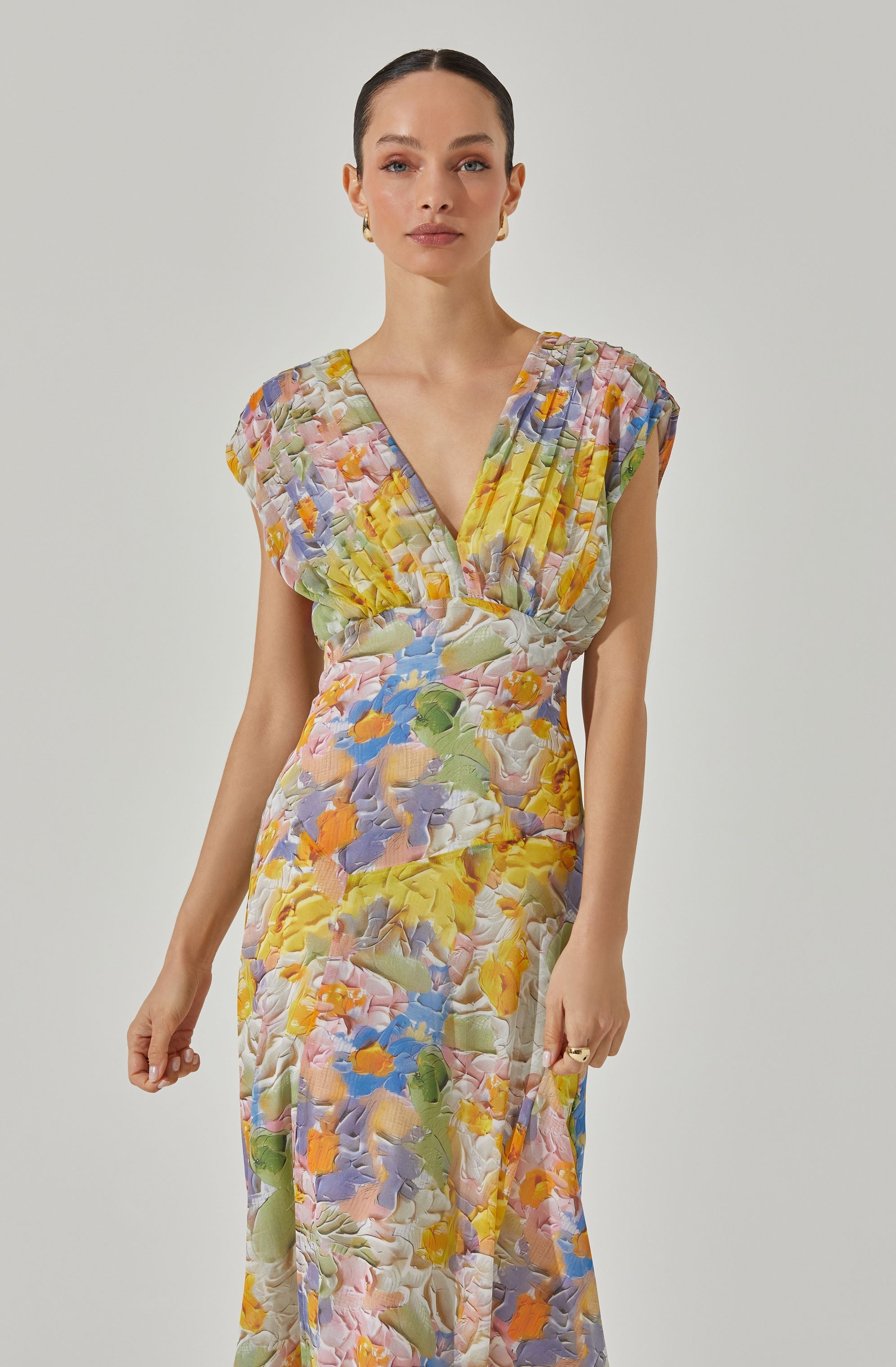 A woman is wearing a colorful, floral-patterned dress featuring a mixture of pastel and vibrant shades. The dress has a V-neckline, with a pleated and ruched bodice that creates a loose fit at the top. The waist is fitted, and the dress flows down in a long skirt with the same floral pattern, giving it a soft, flowing appearance.