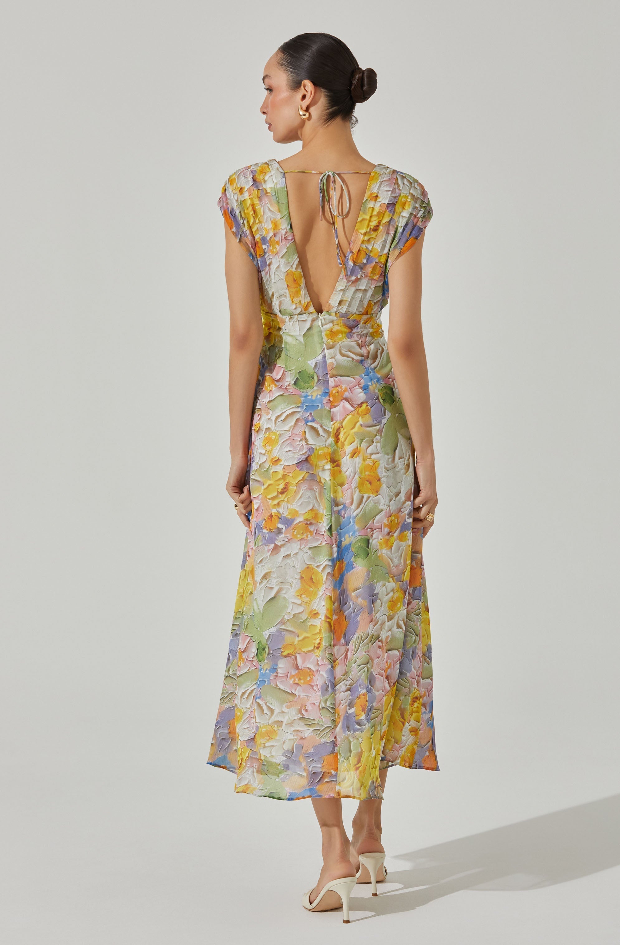 A woman is wearing a long, colorful dress with a floral pattern in pastel shades of yellow, pink, purple, green, and blue. The dress has short, slightly puffed sleeves, a deep V-open back with a tie at the top, and appears to be made of a textured, semi-sheer fabric. She is also wearing a pair of white high-heeled shoes.