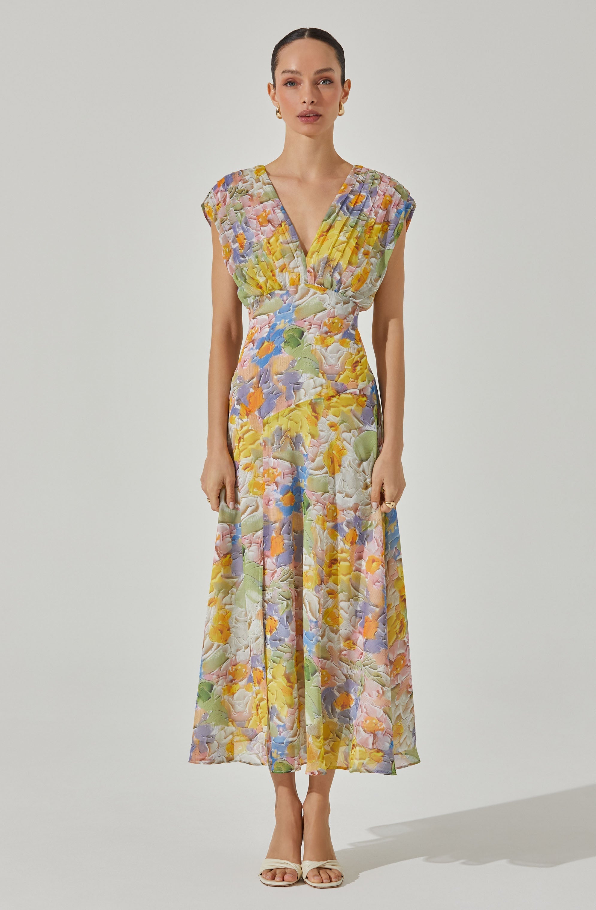 A woman is wearing a sleeveless, ankle-length dress with a V-neckline. The dress features a colorful, abstract floral pattern with pastel shades of yellow, pink, purple, blue, green, and white. The fabric appears lightweight and has a textured, quilted look. She is also wearing nude open-toe heels and gold earrings.