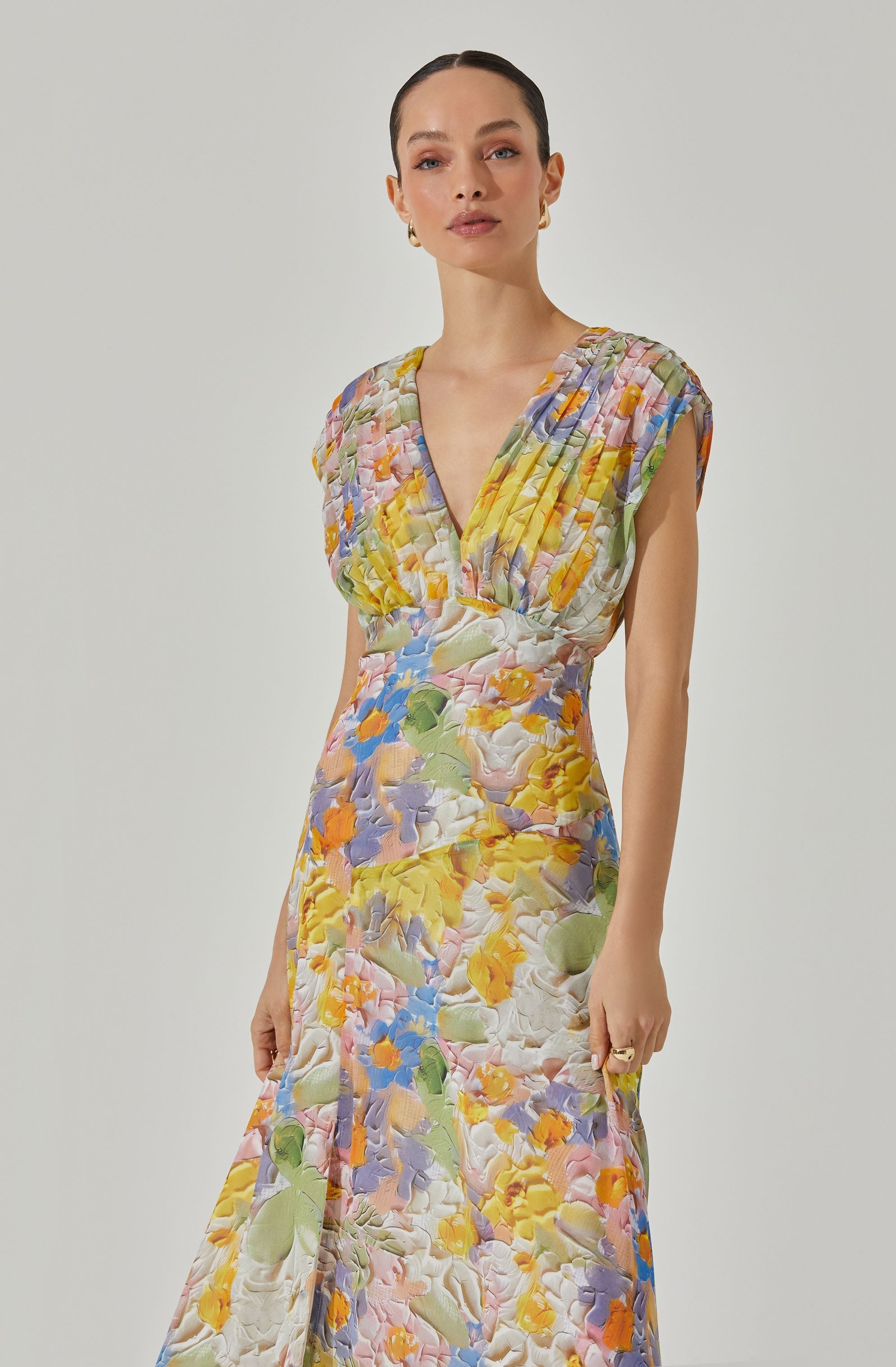 A woman is wearing a sleeveless, V-neck maxi dress with a colorful floral pattern. The dress features a mix of pastel and bright colors, including yellow, pink, purple, blue, green, and white, with gathered detailing at the shoulders and a fitted waist.